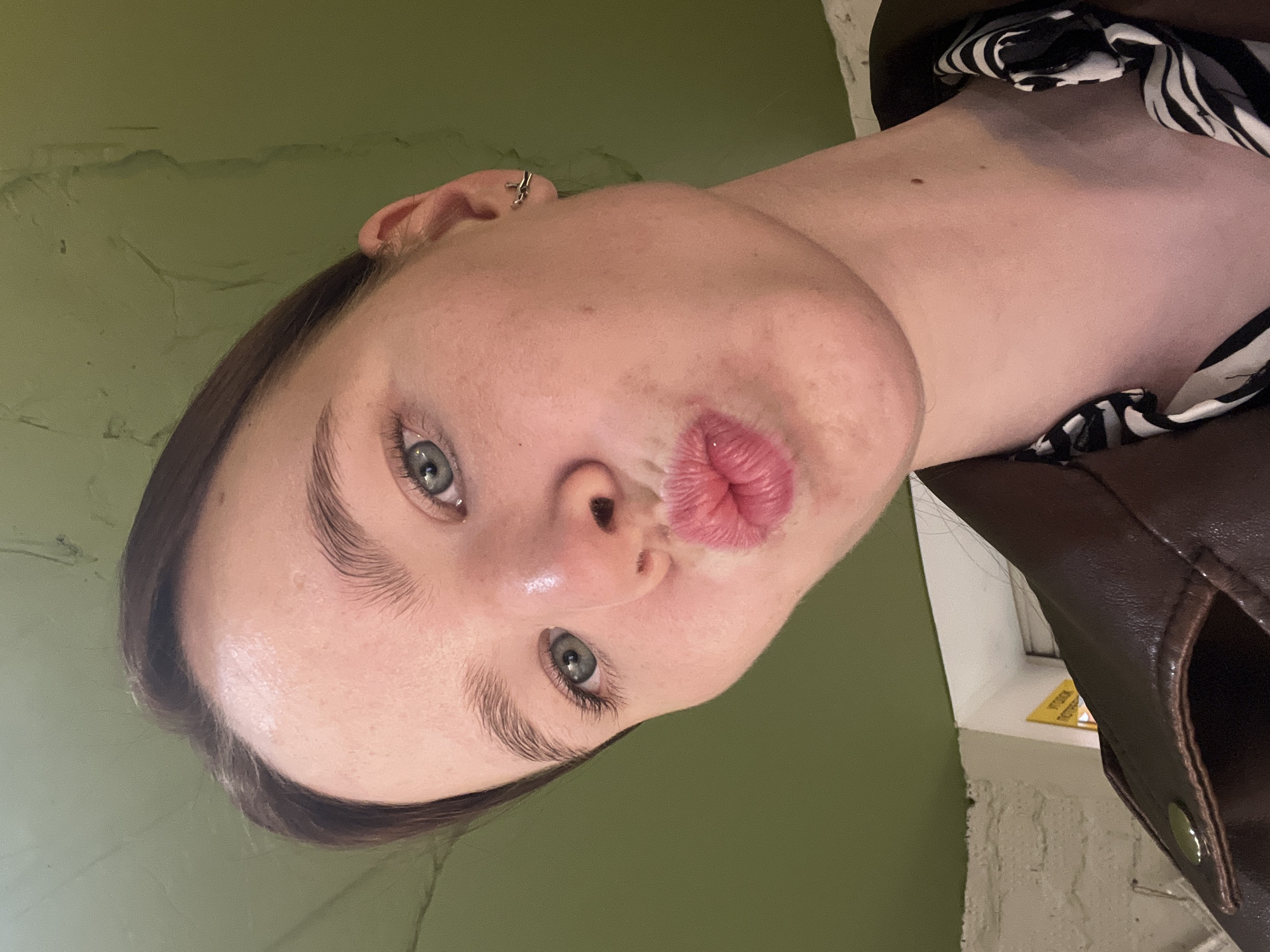 The image is a close-up, rotated portrait of a person indoors, captured from an unconventional upward and tilted angle. The subject, appearing to be a young individual with fair skin, dark hair, and light blue or green eyes, is making a playful "kiss" face with pursed, pink lips. A small silver hoop or stud earring is visible in the upper cartilage of their right ear.
The person is wearing a dark brown or black garment, possibly a jacket or coat, with a hint of a black and white striped pattern visible around the neckline or shoulder. The background is dominated by a muted, olive-green wall, which shows some texture and subtle variations in tone. To the right of the subject, a portion of a window frame or ledge is visible, with a lighter-colored, possibly brick or textured, wall below it. A small yellow object with unreadable dark text sits on the ledge.
The lighting appears to be indoor and uniform, not indicating a specific time of day. The overall impression is one of a casual, lighthearted moment, possibly a selfie. The location context for this image is Chaikovsky, Russia.