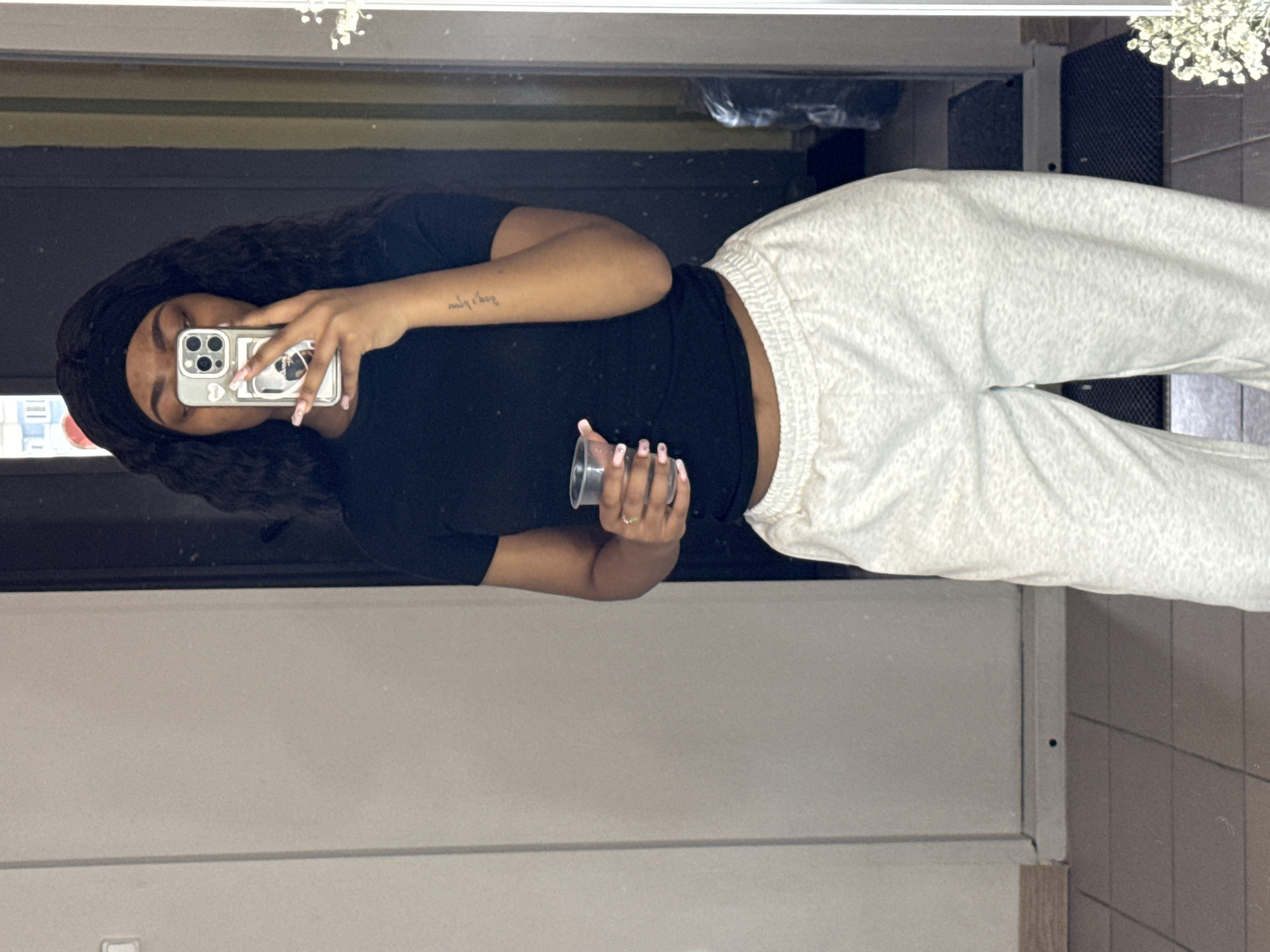A woman stands in front of a mirror, taking a picture of herself with her phone. She has on a black shirt, gray sweatpants, and a black headband, and she is holding a clear plastic cup in her right hand. She is likely inside a building, as the background shows a wall and a cabinet. There are no clear indications of location within Troitsk, Russia.