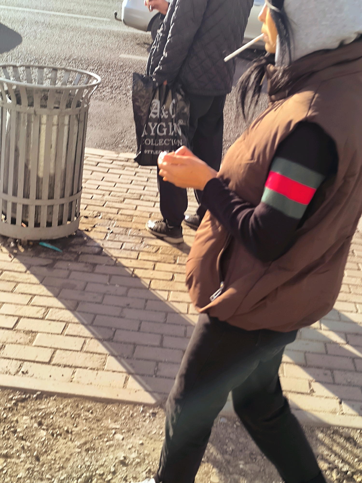 A person stands next to a metal trash can on a sidewalk made of brick pavers. The person is wearing a brown vest over a black long-sleeved shirt with green and red stripes on the left sleeve, black pants, and a grey hooded jacket. They are smoking a cigarette. In the background, another person in a quilted black jacket is visible, as is a white car and a black shopping bag. The sun casts long shadows across the scene.