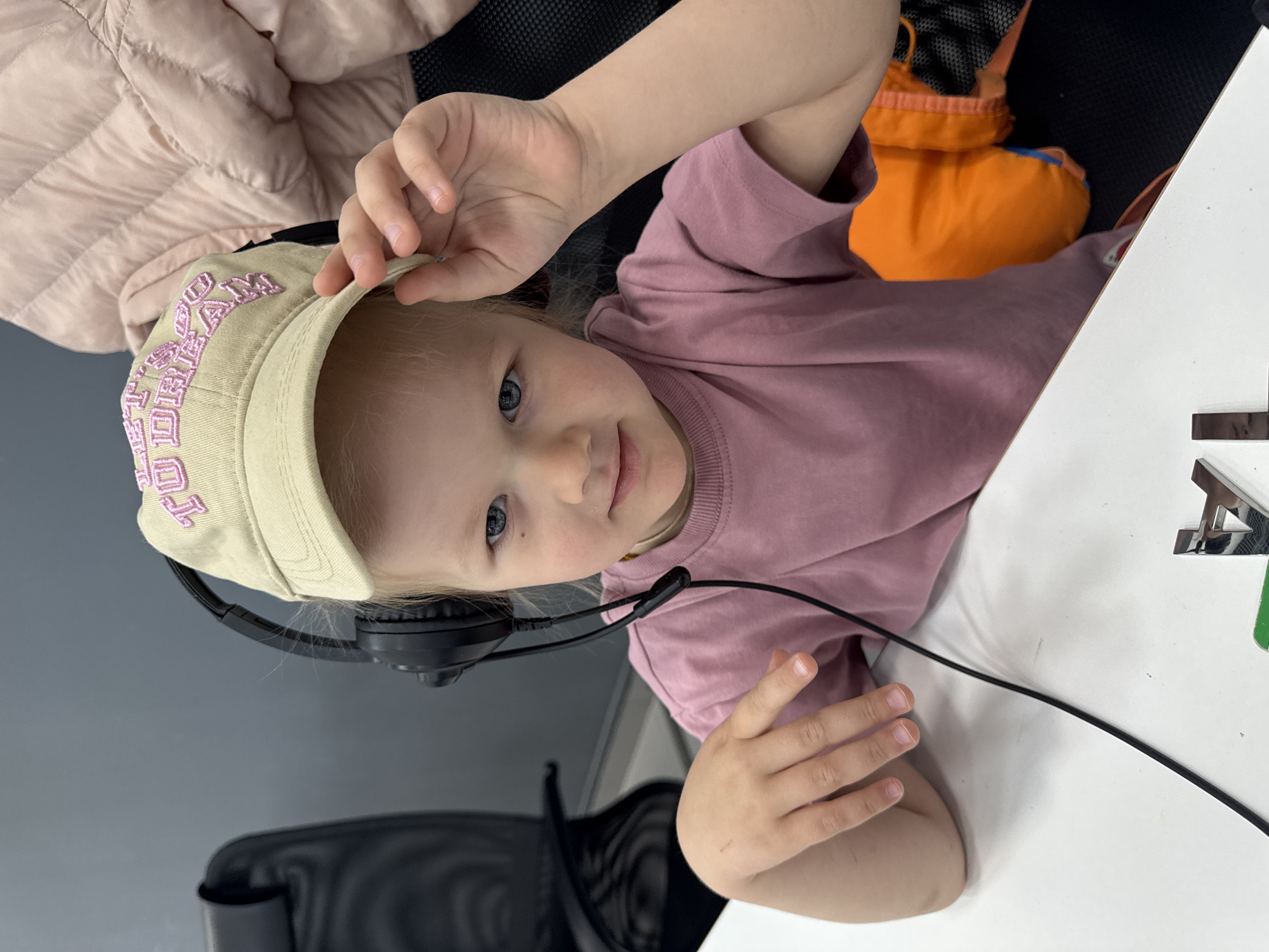 A young child with fair skin and blonde hair is sitting at a white desk while wearing headphones and a beige baseball cap with pink embroidery. The child is looking directly at the camera. The child is wearing a pink t-shirt. An orange bag is behind the child. A black office chair is visible behind the child. There is a peach jacket on the chair. The location is Perm, Russia.