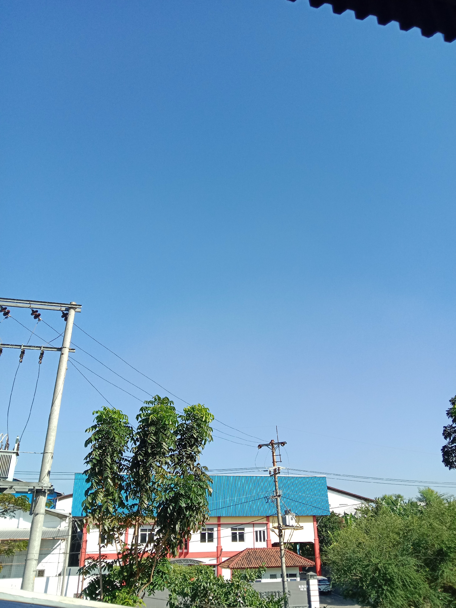The image captures a clear blue sky over a residential or industrial area in Semarang, Indonesia. In the foreground, a utility pole with multiple wires is visible on the left, supporting electrical infrastructure. To the right of the pole, several leafy green trees stand tall. Behind the trees, a building with a prominent blue corrugated roof and white and red walls is situated. Additional structures and more greenery can be seen in the background. The overall scene suggests a typical day in a city with visible power lines and buildings integrated with natural elements.