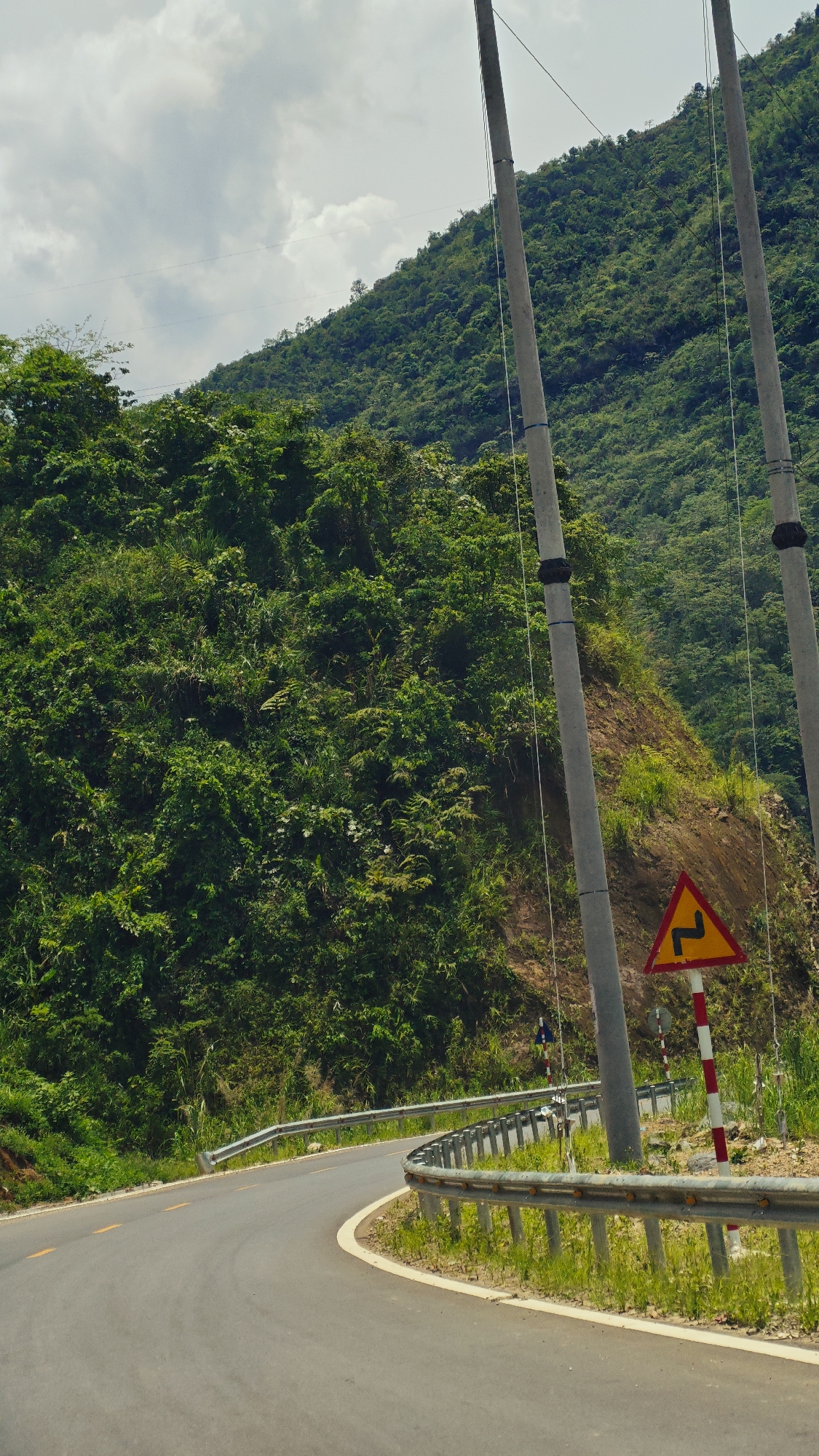 This image depicts a winding mountain road cutting through a densely forested landscape in Duong Quy, Vietnam, under an overcast sky. The scene is dominated by lush green mountains, covered in thick vegetation, that rise steeply in the background and along the left side of the frame.
In the foreground, a paved asphalt road curves sharply to the left. Faint yellow double lines are visible in the center of the road, with a solid white line marking its right edge. A silver-colored metal guardrail runs along the right side of the road, following its curve and separating it from a dirt and grassy bank that ascends into the mountain.
Two tall, grey utility poles stand prominently on the right side of the image, extending vertically with thin wires faintly visible against the green mountain. Adjacent to the guardrail, a yellow triangular warning sign with a red border is displayed on a red and white striped pole. The sign features a black zigzag symbol, indicating a double curve with the first bend to the left.
The sky is a light grey with scattered clouds, indicating an overcast daytime. No specific activities or people are visible, emphasizing the natural, rugged beauty of the mountainous terrain and the infrastructure built within it. The image conveys a sense of a journey through a remote and scenic natural environment.