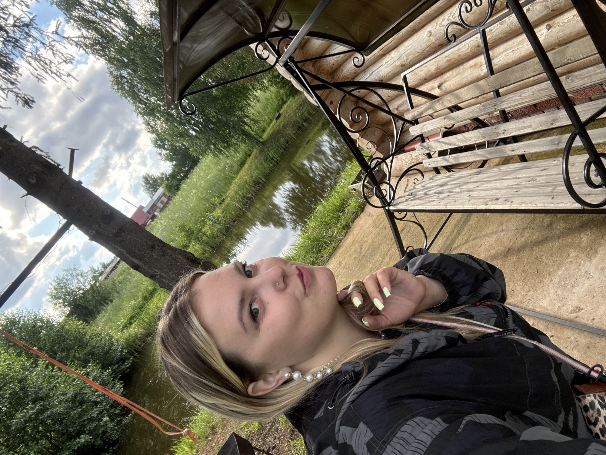This is a close-up, rotated selfie of a young woman with blonde hair and light skin, taken outdoors in what appears to be a rural setting in Markovo, Russia. She is wearing a black jacket with a grey camouflage pattern and a long pearl necklace. Her fingernails are painted a light green color. The woman is looking upwards and to her left, with a slight smile, appearing relaxed and content.
Behind her, the scene includes a body of water, likely a pond or slow-moving river, with lush green grass and trees lining its banks. The sky is cloudy with patches of blue, suggesting it is daytime, possibly late afternoon. To the right of the woman, a wooden structure resembling a gazebo or a porch with a slatted wooden bench and decorative metalwork is visible. The log walls of a building can be seen behind the bench. The overall atmosphere is peaceful and serene, indicating a leisurely moment spent outdoors. No visible text is present in the image.