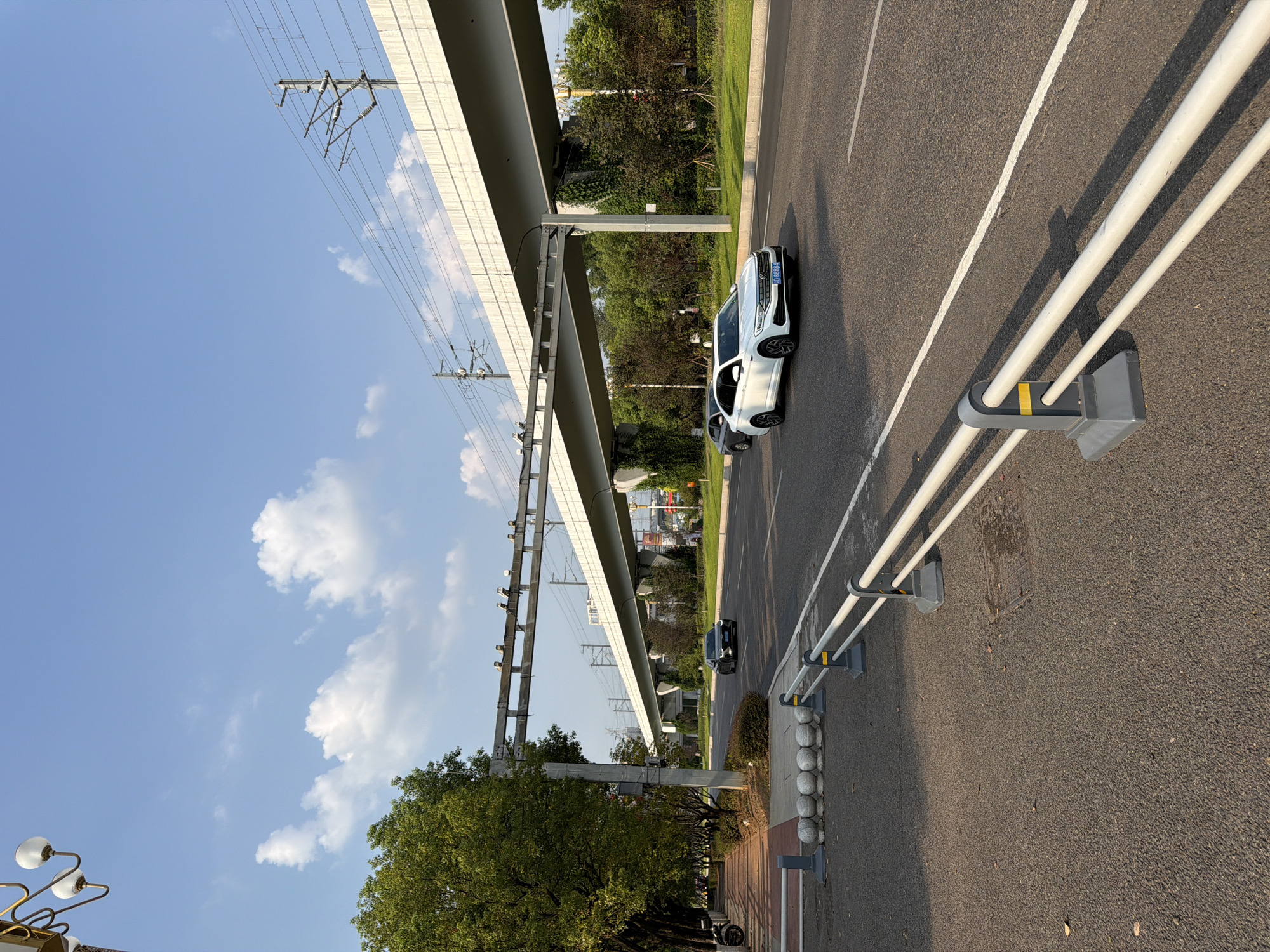The image shows a street scene with a highway overpass and overhead railway lines in Jiangjin, China. A white sedan is driving on the right lane of the road, with a dark sedan behind it. A pedestrian walkway with trees lines the left side of the road. In the background, lush green trees and vegetation are visible. The sky is clear with a few scattered clouds. The street appears to be a major thoroughfare, with the overpass suggesting a significant transportation hub. The presence of both cars and railway lines indicates a complex transportation infrastructure. The scene is captured during daylight hours, likely in the late afternoon due to the long shadows cast by the guardrail.