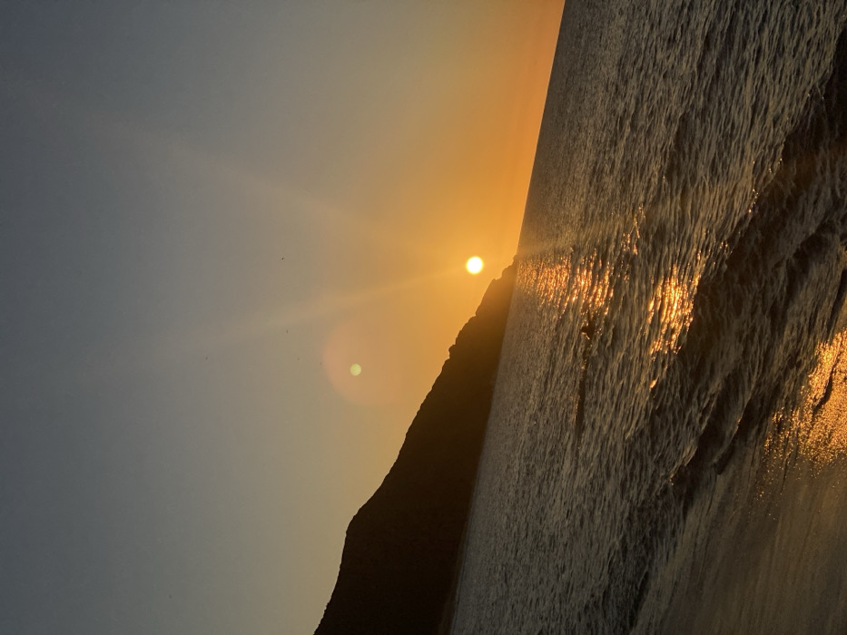 The image displays a coastal landscape during either sunrise or sunset, with the sun low on the horizon. The scene is set on a coastline, identified as being in the city of Altagracia, Venezuela.
The sun, a distinct, bright orange sphere, is positioned above the water towards the center-right of the frame, emitting visible golden rays that extend upwards into the sky. The sky transitions from a deep blue-grey at the top to vibrant orange and yellow tones near the horizon. A prominent, circular greenish-yellow lens flare artifact is present in the upper-left portion of the sky. Several small, dark, indistinct specks, consistent with distant birds, are visible against the sky in the upper-left quadrant.
A dark, silhouetted mountain or significant hillside occupies the left side of the frame, extending from the left edge and sloping down towards the center-right, partially obscuring the sun's base. The water, likely ocean or sea, covers the middle ground, appearing dark with undulating waves. A shimmering path of intense golden-orange light reflects off the water's surface, extending from the sun towards the foreground. In the lower-left corner, a wet sandy or rocky shoreline is visible, also reflecting the golden light from the sun. The horizon line runs diagonally from the lower-left to the upper-right. No human figures, man-made structures, or explicit actions are discernible within the frame.