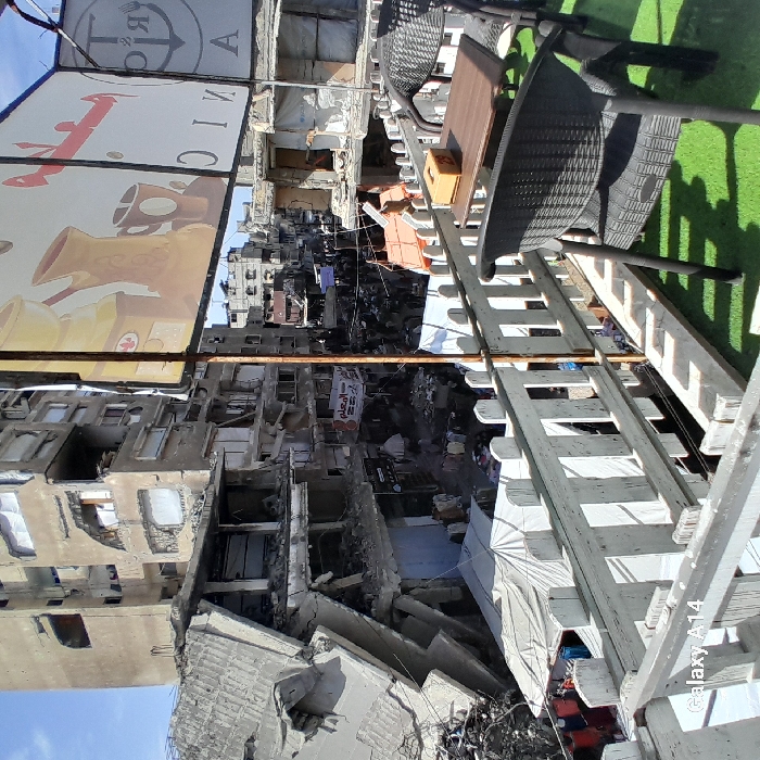 This image depicts a scene of extensive urban destruction in Khan Yunis, Palestinian Territory, viewed from an elevated perspective, likely a balcony or rooftop.
In the immediate foreground, a white wooden picket-style railing spans horizontally, partially obscuring the view downwards. The floor within this foreground space is covered with green artificial turf. Two dark-colored woven chairs are positioned, one fully visible on the right, and a small wooden table with a tissue box rests between them. Above this area, a large rectangular sign, partially visible, displays images of a coffee grinder, a traditional jug, a coffee cup, and Arabic text alongside an anchor symbol, suggesting a cafe or similar establishment. A thin metal pole supports this sign, extending vertically near the center-right.
Below the railing, the midground reveals a street or market level characterized by widespread debris and a dense concentration of people. Numerous individuals, appearing as civilians, are visible moving and standing amongst rubble and makeshift market stalls. Several temporary white canvas tents or tarpaulins are erected on the ground, some sheltering merchandise or groups of people. Goods appear to be laid out on the ground for sale.
The background is dominated by severely damaged multi-story buildings. On the left, a tall concrete structure shows catastrophic collapse on its lower and middle floors, with large sections of concrete detached, floors caved in, and exposed rebar. Another multi-story building further back also exhibits significant structural compromise, with broken windows and visible interior damage. Buildings lining the street on the right are similarly affected, featuring large openings where walls or floors have collapsed. A banner with Arabic text is visible on a pole or building in the mid-background. The sky above is clear and blue.