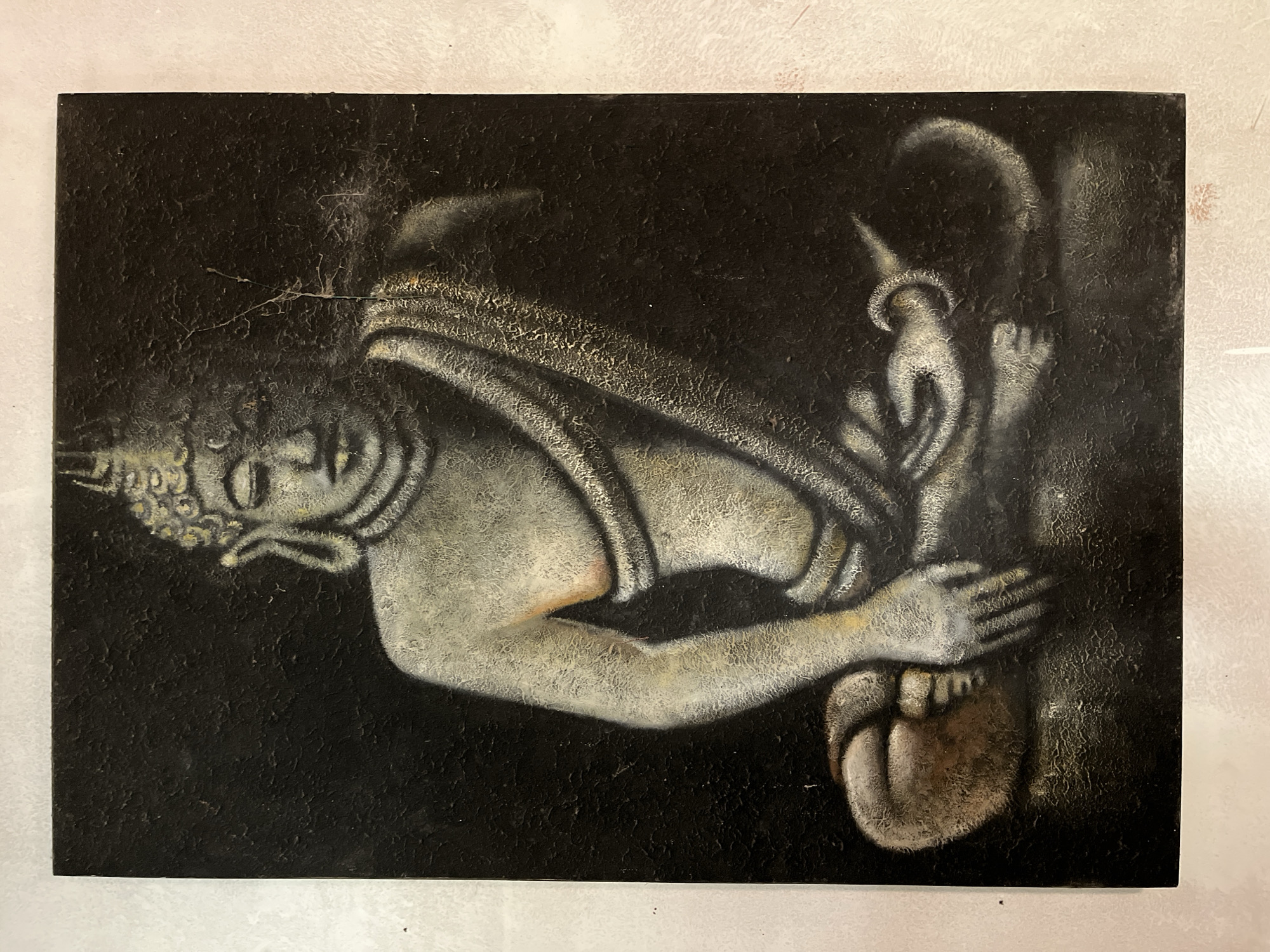 A painting depicts a seated figure, likely Buddha, in meditation. The figure is rendered in shades of grey and white against a dark, textured background. The subject's face is serene, with closed eyes and a crown-like headdress. Draped robes cover the body, with one arm resting on the knee and the other extended. The lower body is crossed in a meditative pose. The painting exhibits a rough, textured surface, possibly achieved through impasto techniques or a heavily textured canvas. The overall mood is contemplative and spiritual. The painting is located in a building in Karangkebagusan, Indonesia, though no specific details about the surrounding environment within Karangkebagusan are visible in the image itself.