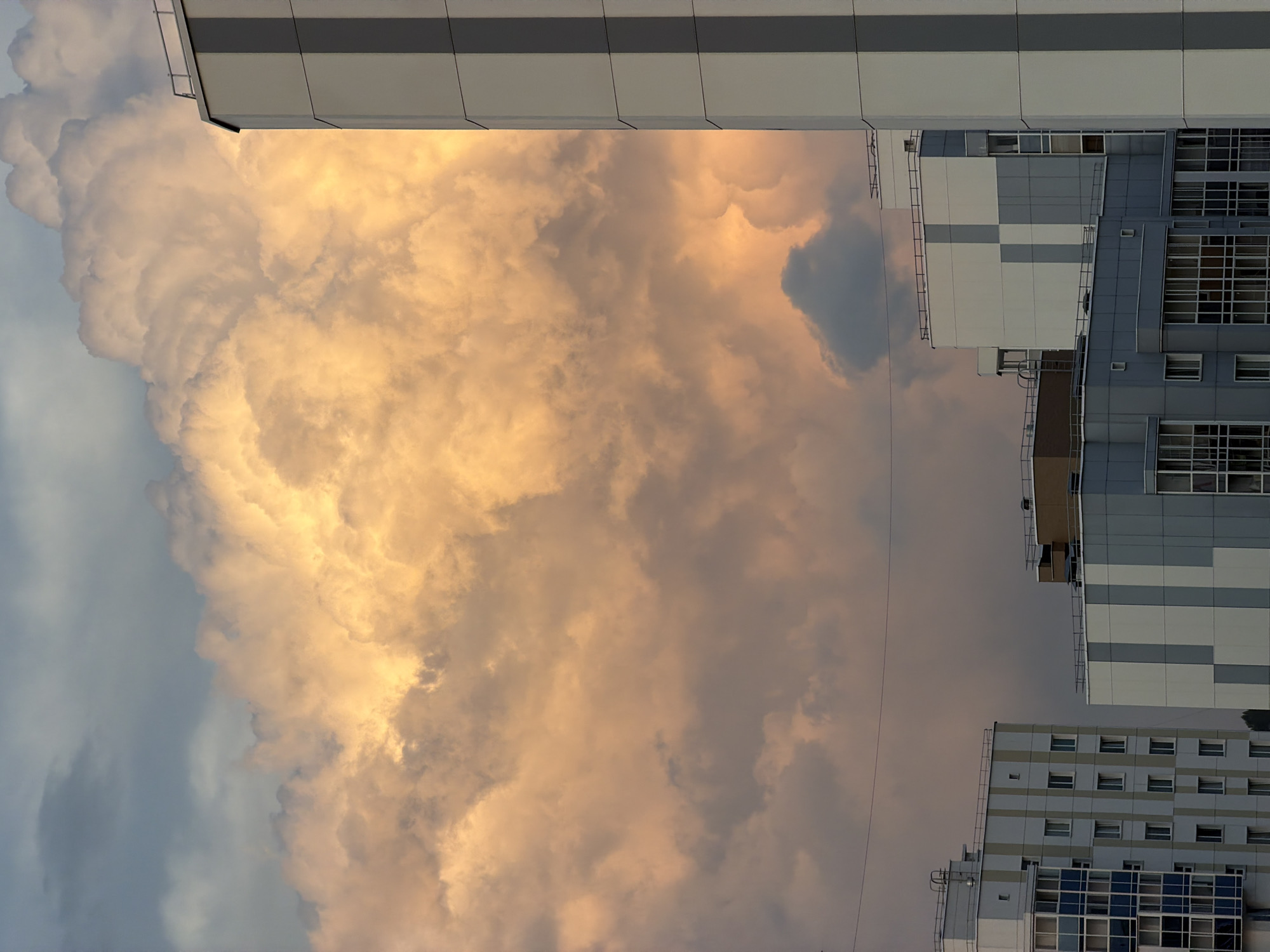 The image shows a sky filled with dramatic, illuminated clouds. The clouds are a mix of golden-orange and gray tones, with the golden-orange clouds appearing to be lit from behind. In the foreground, there are several modern apartment buildings. The building on the far right is tall and predominantly white with vertical gray stripes. To the left of it, other apartment buildings of varying heights and designs are visible, featuring white, gray, and blue facades with numerous windows. A thin wire runs horizontally across the upper portion of the image, and there are some faint details of rooftop structures on the buildings. The overall scene captures a dynamic sky over an urban residential area, likely in Tsiolkovsky, Russia, given the architectural style of the buildings. There are no people or animals visible in the image.