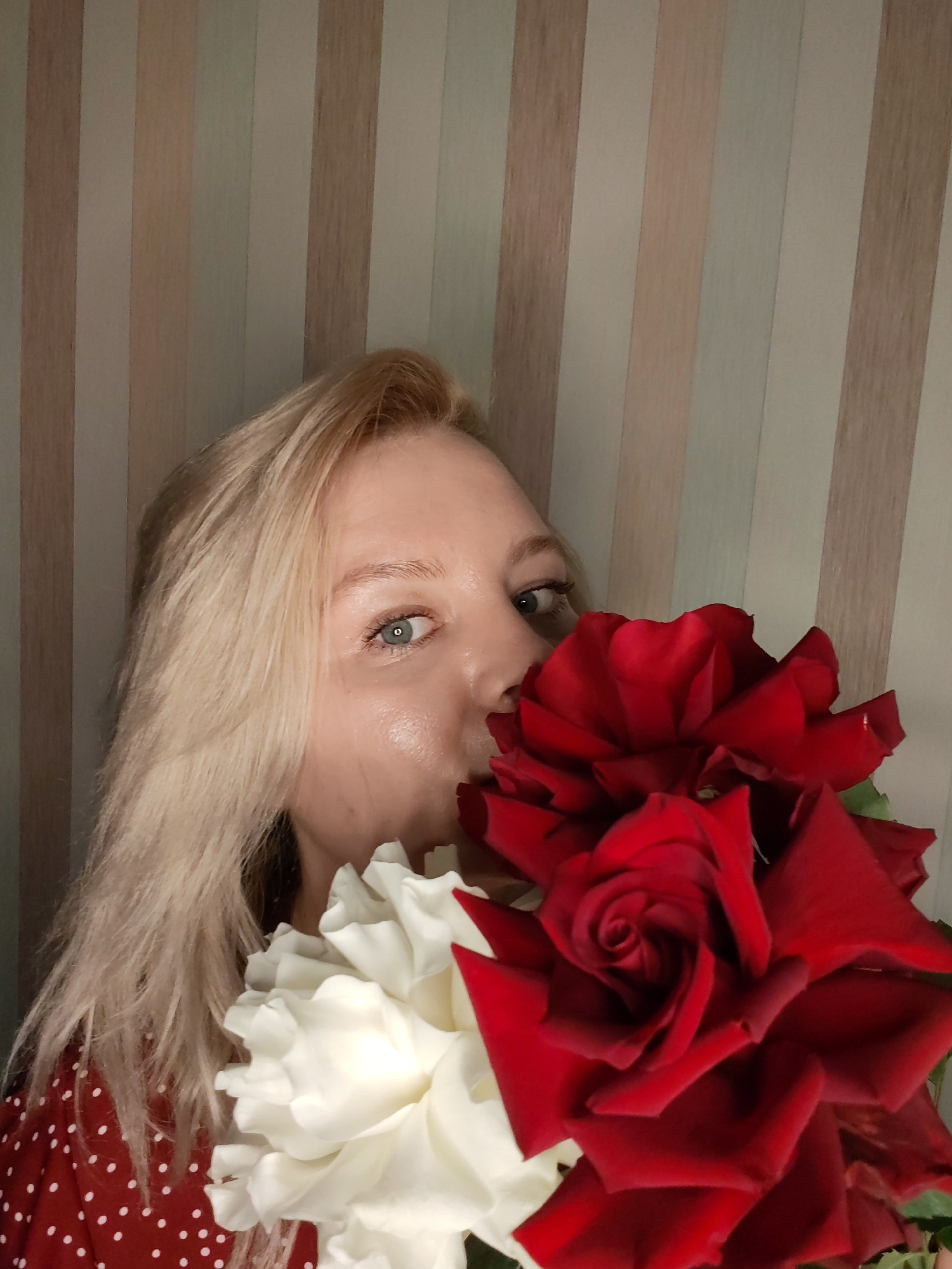 An image captures a blonde woman holding a large bouquet of roses, primarily red and white, up to her face. Her left blue eye is visible, looking directly at the camera, while the rest of her face is mostly obscured by the flowers. Her fair skin and blonde hair are prominent. She is wearing a red garment with white polka dots, visible on her left shoulder and arm. The background consists of a wall with vertical stripes in muted tones of beige, cream, and light brown, suggesting wallpaper in an indoor setting. The lighting is soft and even, not indicating a specific time of day. The woman appears to be smelling the roses or playfully hiding behind them, conveying a gentle or appreciative mood. There is no visible text in the image. The location context for the image is Izhevsk, Russia.