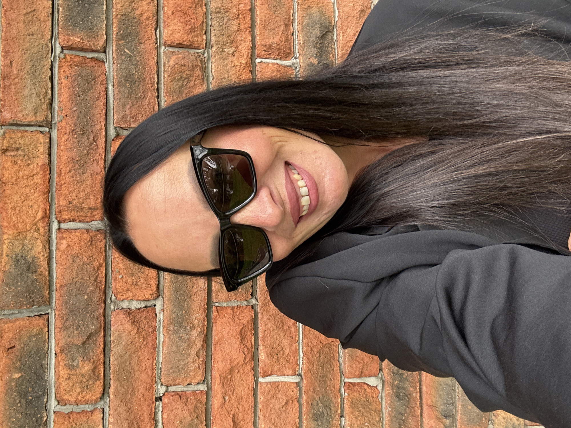 A woman with long, dark hair and sunglasses smiles against a brick wall. She is wearing a black blazer. The image is a selfie taken outdoors.