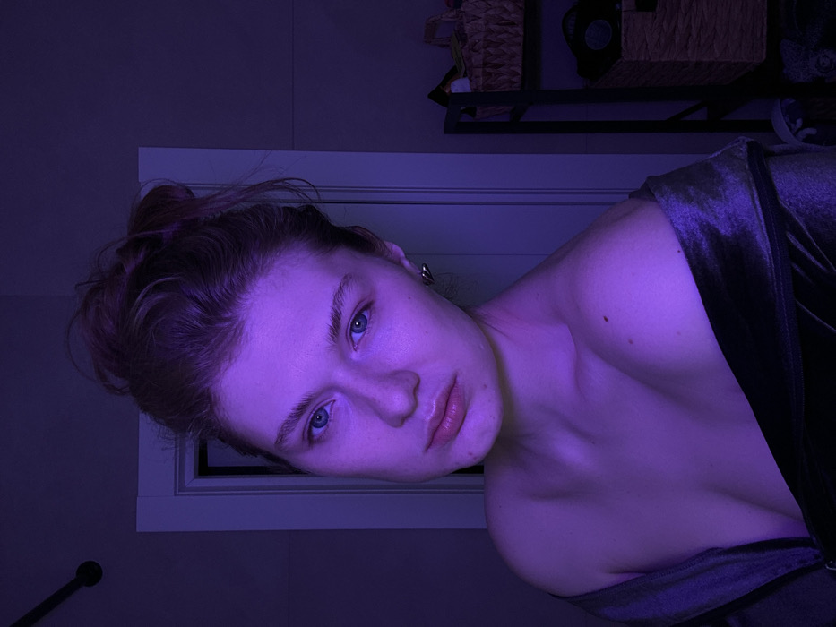 A woman with fair skin is angled to the left, gazing at the camera under violet lighting. Her auburn hair is gathered in a loose bun. She has blue eyes, full lips, and a mole on her chest. A velvet fabric drapes off her shoulders. Behind her is a closed white doorframe and a shelf with wicker baskets and dark objects. A slender black rod is visible in the lower left corner.