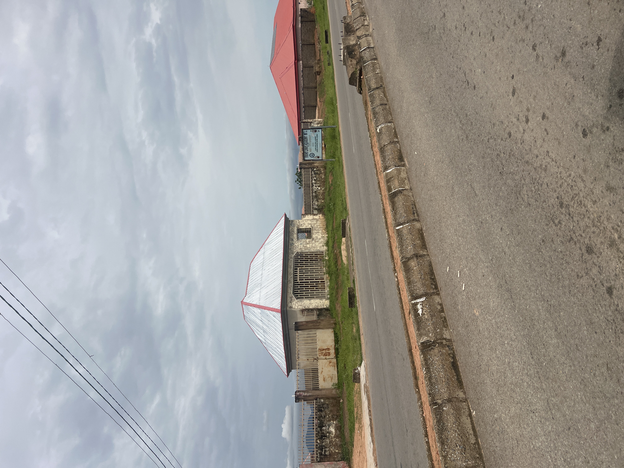 The image shows a street scene in Kona Garu, Nigeria. The perspective is from a slightly elevated angle, looking down towards the road and adjacent buildings. The sky is overcast with fluffy white clouds, suggesting a daytime scene.
On the left side of the image, there are power lines running diagonally across the frame. Further to the left, partially obscured by the power lines, are more buildings, one with a distinctive red roof. In the foreground, there is a building with a white and red corrugated metal roof, constructed with stone walls. It appears to be a residential or commercial building, with barred windows and a metal gate. Behind this building, a sign is visible, attached to a fence. The text on the sign reads "FOUNDATION ACADEMY INT'L." The surrounding area is covered with grass.
The road occupies the right side of the image and is paved with asphalt. It is bordered by a raised kerb made of concrete blocks. The road surface shows some signs of wear and tear, with darker patches and a few small white objects that might be litter.
The overall impression is a typical street view in a Nigerian town, with a mix of residential and possibly educational buildings along a main road. The cloudy weather adds a muted quality to the scene. There are no people visible in the image.