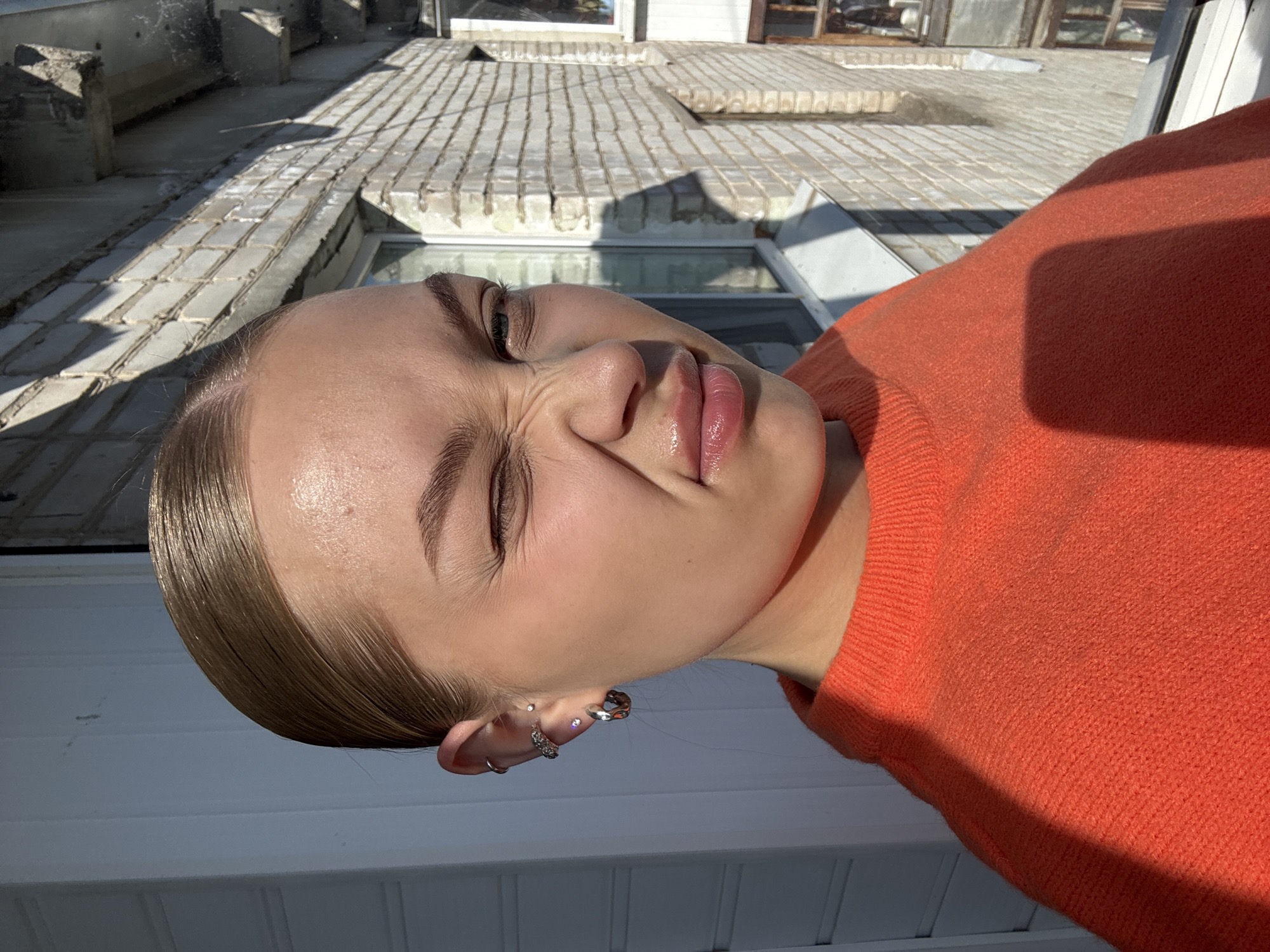 A young woman with fair skin and light brown hair slicked back is pictured close-up, with an orange sweater visible. Her eyes are squinted shut, and her face is contorted in a grimace. Multiple silver earrings are visible in her left ear. The background is a building facade of light-colored brick and what appears to be windows with light-colored frames. It is daytime and the lighting appears to be natural sunlight. The location is likely Chaikovsky, Russia.