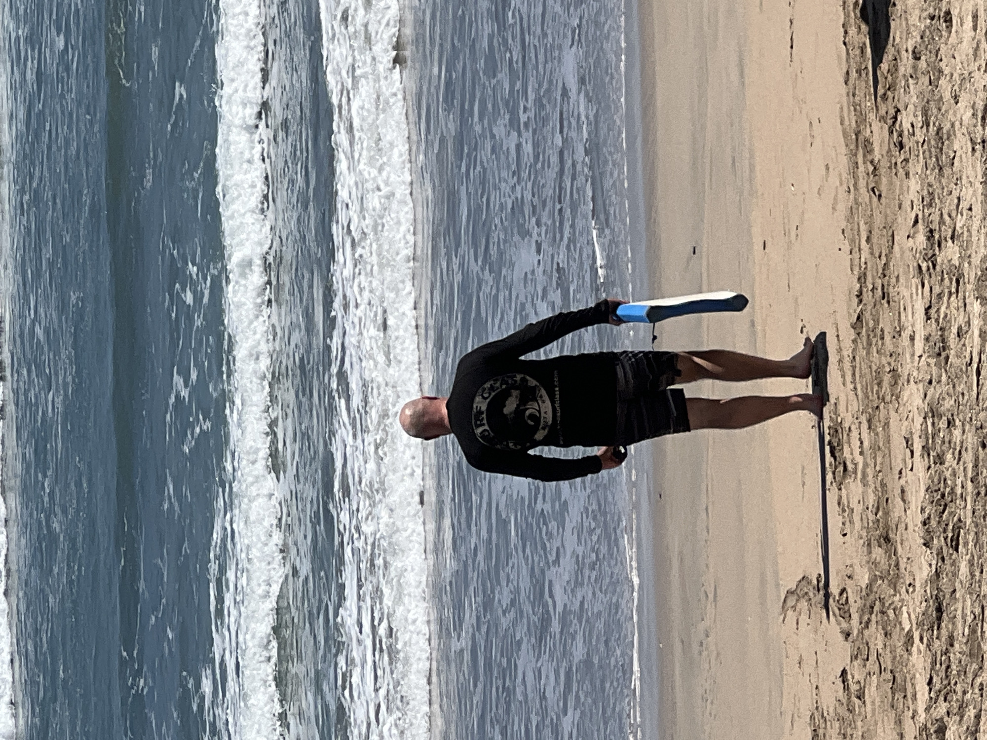 A bald man wearing a black long-sleeved shirt and striped swim trunks stands on a sandy beach, facing the ocean. He is holding a blue and white boogie board. The ocean has moderate waves with white foam. The sand in the foreground is wet, indicating the tide is out or receding. The background shows more waves and the horizon. The scene is likely Kuta Beach in Bali, Indonesia, known for its surf.