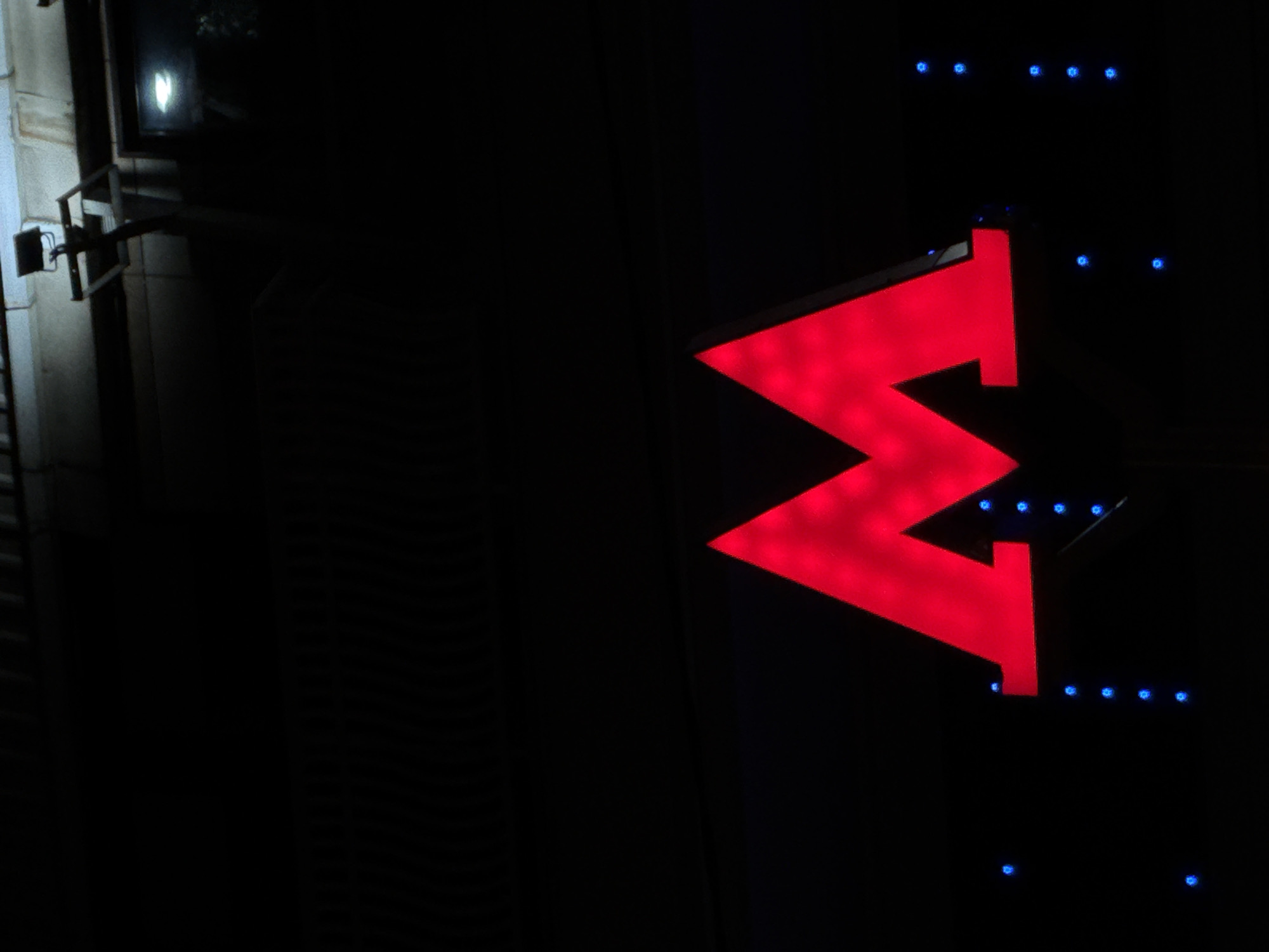 The image shows a red "M" sign, illuminated with small blue lights, indicating the Moscow Metro entrance at night. The sign is mounted on a dark building. To the left is a wall and metal framework. The background is black, suggesting it is dark.