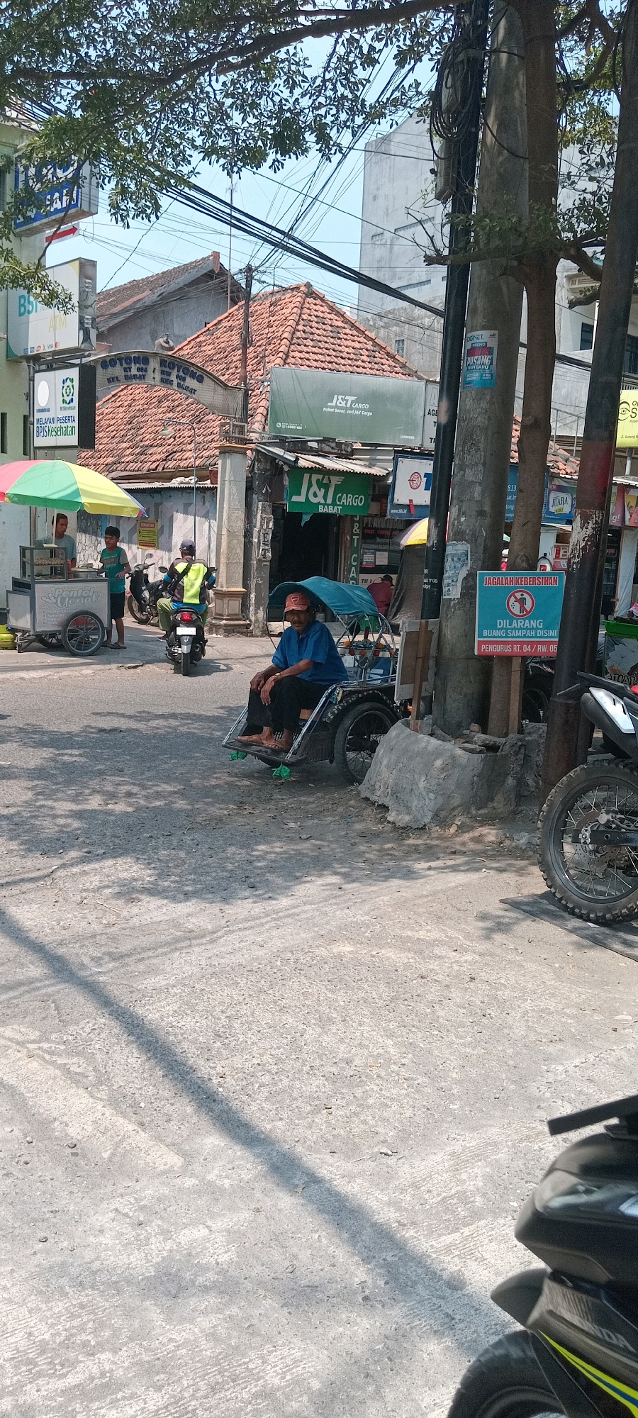 The image shows a street scene in Babat, Indonesia. In the foreground, a person wearing a blue shirt and a red cap is seated in a three-wheeled vehicle, possibly a pedicab or becak, with a blue canopy. The vehicle has two large rear wheels and a smaller front wheel. The person appears to be resting. Behind the seated person, a motorcycle with a rider wearing a bright yellow vest is visible. To the left, there is a food cart under a colorful umbrella, with two young males standing near it. The cart has text that reads "Pentol Uenak" which translates to "Delicious Pentol". Further back, there is a building with a terracotta tiled roof. A sign for "J&T Cargo" is prominently displayed, indicating a logistics service. Another sign with the text "GOTONG ROYONG" and addresses suggests a local community initiative or business. Electrical wires are strung across poles, common in urban environments. A sign with a red circle and a diagonal line through a trash can symbol, along with text that reads "JAGALAH KEBERSIHAN" and "DILARANG BUANG SAMPAH DI SINI" (Keep Clean and Do Not Litter Here), is posted on a pole. The ground is paved with asphalt and concrete, with shadows from trees and overhead wires casting patterns. A motorcycle wheel is visible in the lower right corner, suggesting another vehicle present. The overall impression is a busy, sunlit street with commercial activity and everyday life unfolding.