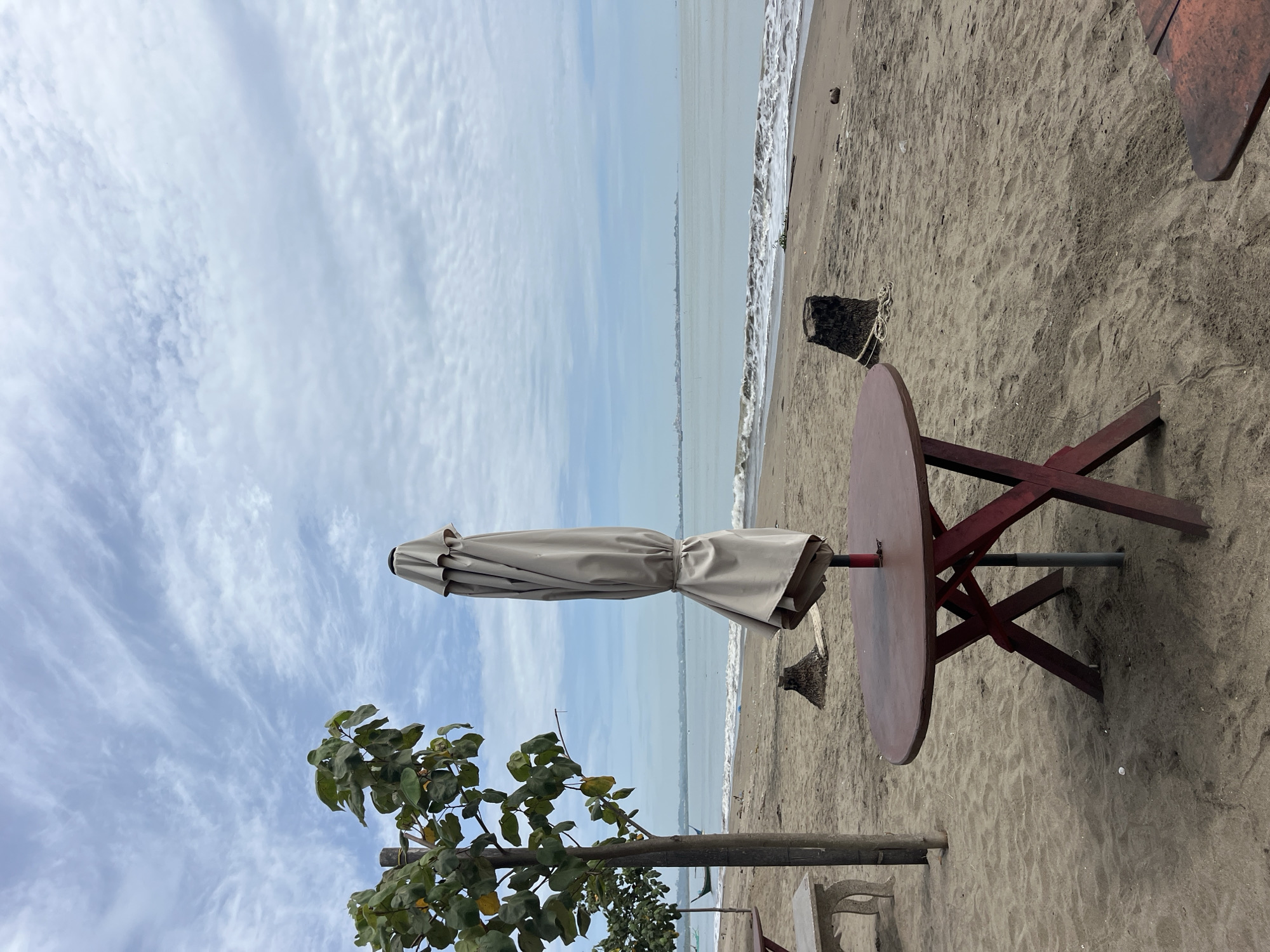 The image depicts a beach scene in Lhokseumawe, Indonesia. The beach is sandy with a partly cloudy sky overhead. A folded beige beach umbrella is attached to a round, dark red table with a red trestle base. The table is set on the sand. In the lower left corner, a tree with green leaves is visible. The ocean with small waves extends to the horizon. A wooden post and a second table can be seen in the foreground, just behind the leaves of the tree.