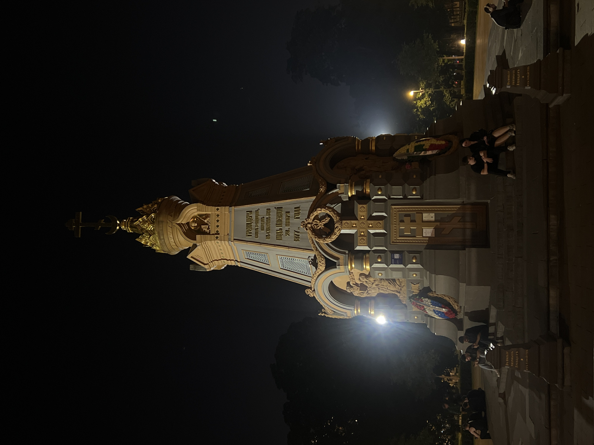 In an urban park at night, a large, ornate monument stands illuminated by spotlights. The monument is made of stone and gold accents, featuring intricate carvings and religious iconography, including a cross and a crescent moon atop its spire. Inscriptions in Cyrillic are visible on its facade. A wreath of greenery and red, white, and blue ribbons adorns the monument's base.
Several individuals are gathered around the monument's base. Two young men sit on steps to the left, looking at their phones. To the right, a couple sits closely together on the steps, with the woman resting her head on the man's shoulder. Another person is seated on the ground to the far right, facing away from the monument.
The background shows dark trees and a few streetlights, suggesting a dimly lit urban environment. The sky is dark with a faint haze.