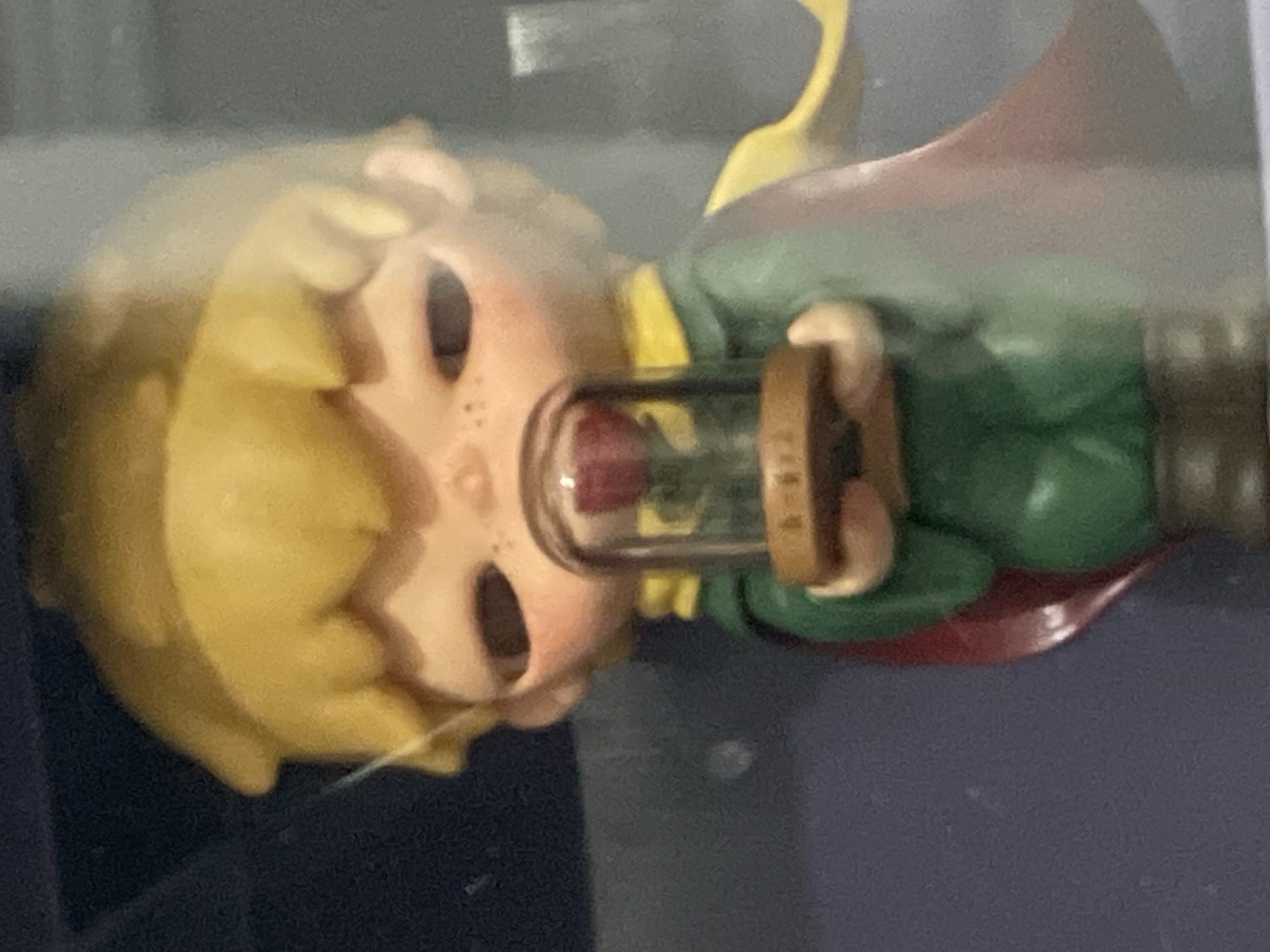This media file is a close-up, vertical photograph of a figurine resembling the character "The Little Prince." The figurine has blonde hair, rosy cheeks with freckles, and large, dark eyes. It is depicted holding a small glass dome containing a red rose. The figurine is dressed in a green tunic, yellow scarf, and a reddish-brown cape. Its pose suggests it is presenting or protecting the rose.
The lighting in the image is somewhat diffused, making it difficult to determine the exact time of day or weather conditions. The background is out of focus, but appears to be a display case or shelf with a dark, possibly blue, surface. There are no clear location cues to definitively place it in Denpasar, Indonesia, beyond the provided context. The overall mood conveyed by the figurine is one of innocence and perhaps a touch of melancholy, consistent with the character's portrayal. No visible text is present in the image.