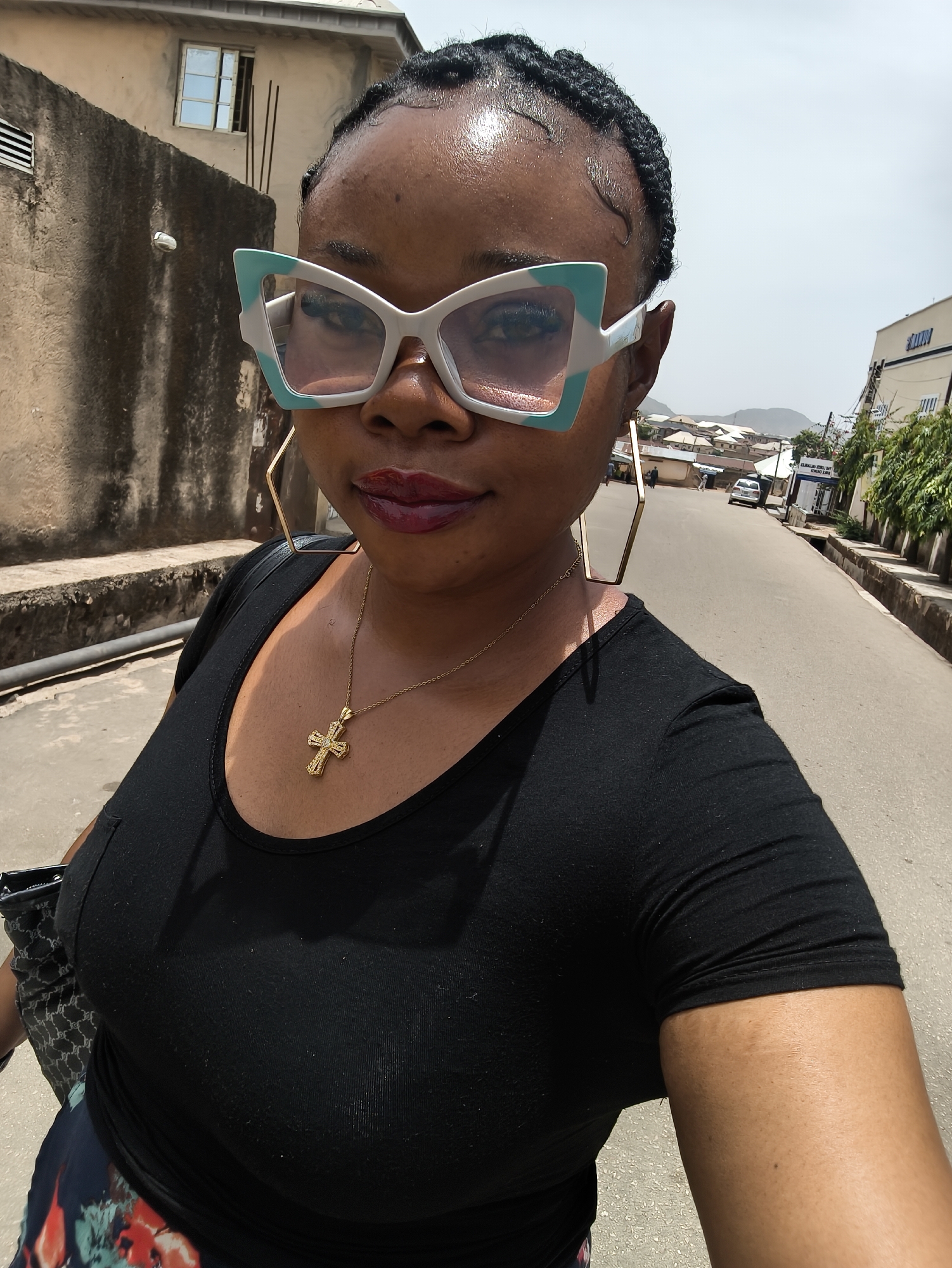 A woman is featured in a close-up selfie, standing on a paved street in Kabong, Nigeria. She has dark skin, with her hair styled in small braids. She wears dark red lipstick and distinctive large cat-eye sunglasses with white frames and turquoise accents. A gold chain necklace with an ornate gold cross pendant rests on her chest, and she has large, geometric gold hoop earrings. Her attire consists of a black, short-sleeved t-shirt and a colorful floral-patterned skirt, partially visible at the bottom left. A black bag is slung over her left shoulder.
The background reveals a typical street scene. Behind her, buildings of various styles and textures line the road, with one beige building featuring a visible window. Further down the street, another building clearly displays the letters "SUN" on its facade. In the distance, low hills or mountains are visible under a bright, clear sky, suggesting sunny midday weather. The road extends into the distance, with a white car and a few individuals visible further along. On the right side of the street, a curb and what appears to be a drainage ditch are present, alongside some sparse green vegetation. The woman appears to be taking a photo of herself while outdoors on the street.