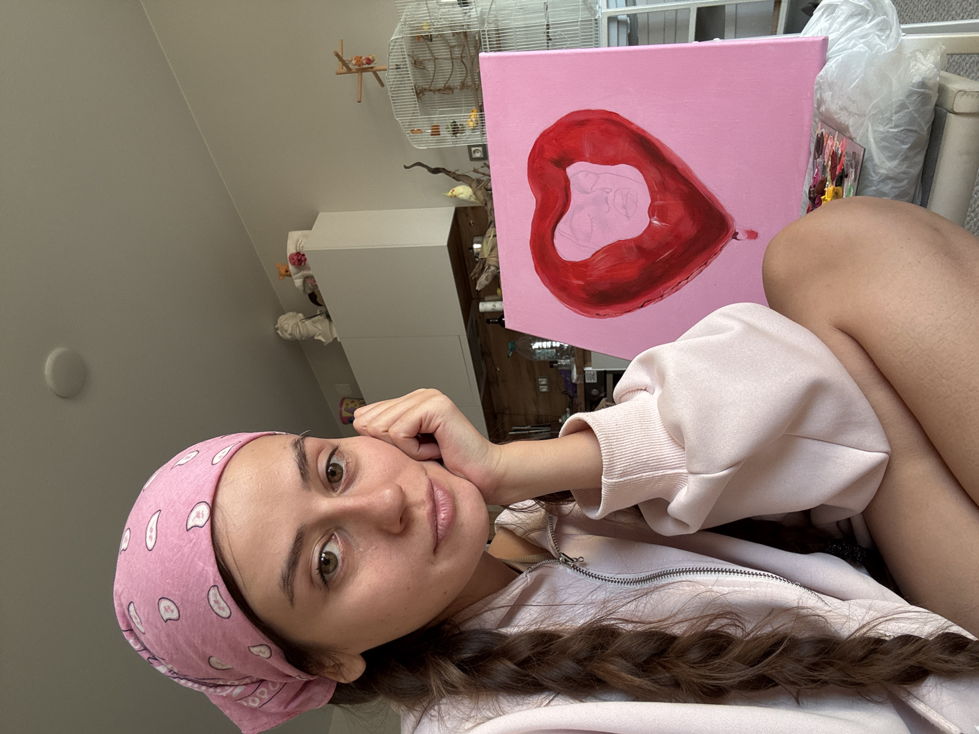 A young woman is captured in a selfie-style portrait, looking directly at the camera with a calm, pleasant expression. She is wearing a pink bandana with white paisley patterns and a light pink zip-up hoodie. Her dark brown hair is styled in a braid, partially visible. Her left hand is resting gently under her chin.
To her right, a canvas painting is prominently displayed, showing a large, glossy red representation of lips on a solid pink background. Inside the red lips, a faint outline of a heart in a lighter pink suggests the artwork is still in progress. Next to the painting, art supplies including a palette with various paint colors and a plastic bag, possibly containing more supplies or brushes, are visible.
In the background, a white birdcage with birds inside can be seen, along with a perch for birds. A yellow bird is perched on a branch outside the cage. The setting appears to be an indoor room with a sloped ceiling, indicating an attic or a room with an architectural feature. White furniture, possibly a cabinet or shelving unit, is also in the background. The lighting suggests it is daytime.
The image portrays a moment of an artist, likely in her home or studio, taking a break or posing with her ongoing work and pets. No visible text is present in the image. The location context provided is Krasnoyarsk, Russia.