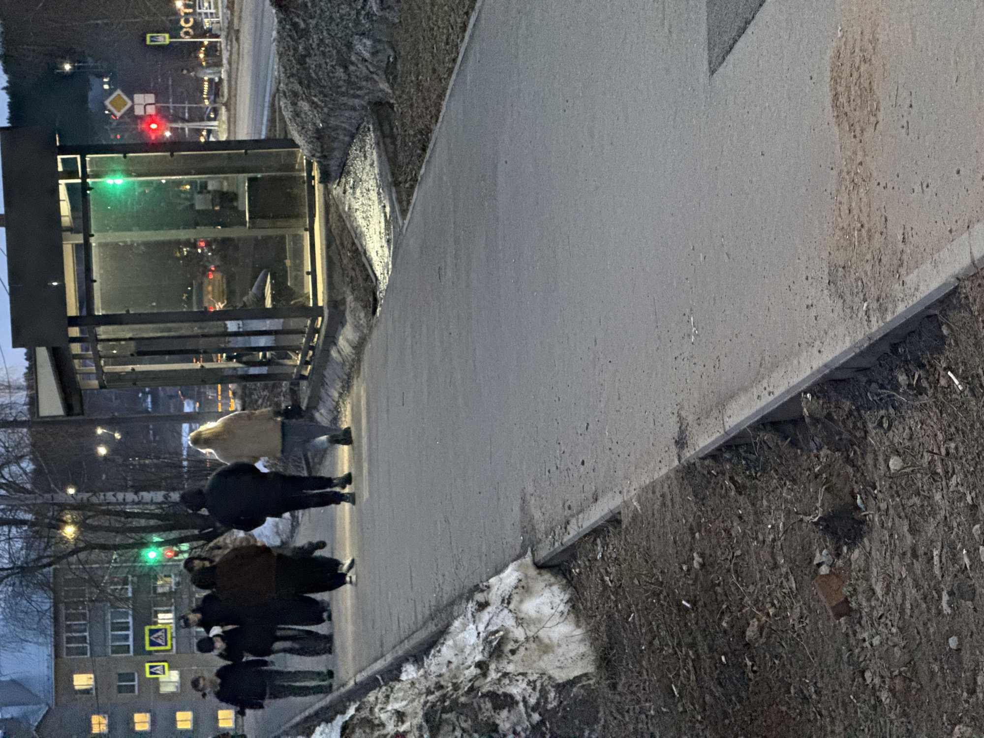 The image depicts an urban scene in Votkinsk, Russia. A group of approximately seven people is walking along a sidewalk. Adjacent to the sidewalk is a patch of ground covered with dirt and remnants of snow. A bus stop shelter is visible on the left. Traffic signals are present; one is red and one is green. A crosswalk sign is visible in the background near an apartment building. The scene appears to be taken during early evening or twilight.