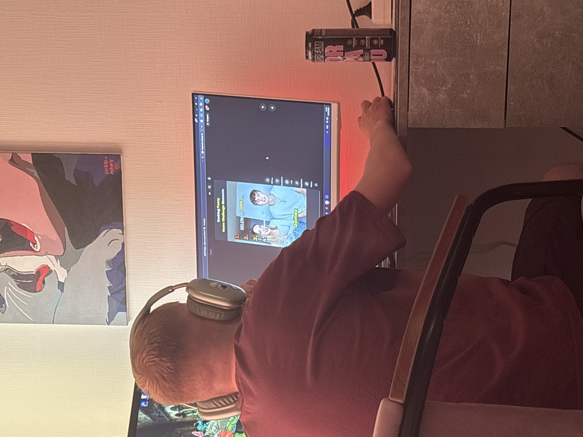 A person is seated at a desk, viewed from behind, wearing a reddish-maroon t-shirt and headphones around their neck. Their attention is directed towards a computer monitor, which displays what appears to be a video from a platform like YouTube. The video shows a ranking or list, with visible text including "Ranking Funny", "Challenge Moments", and "SELENA GOMEZ" and "Gomez" next to an image of a person.
Behind the monitor, a red glow emanates, possibly from LED strip lighting, casting a warm hue on the wall. On the wall to the left, a framed piece of art is visible, featuring a stylized figure in shades of red, black, and grey, with the text "AMSU-MONY" written on it.
To the right of the monitor, an open black and red can, labeled with "CREAM" and "OR 4D", is positioned upright and plugged into a wall outlet. The person's right hand is extended towards the wall outlet area, possibly interacting with the can or the cable. The setting appears to be an indoor room, likely an office or bedroom in Sinyavino, Russia, with the overall ambiance suggesting evening or night due to the artificial lighting. The person seems engaged with the content on the screen.