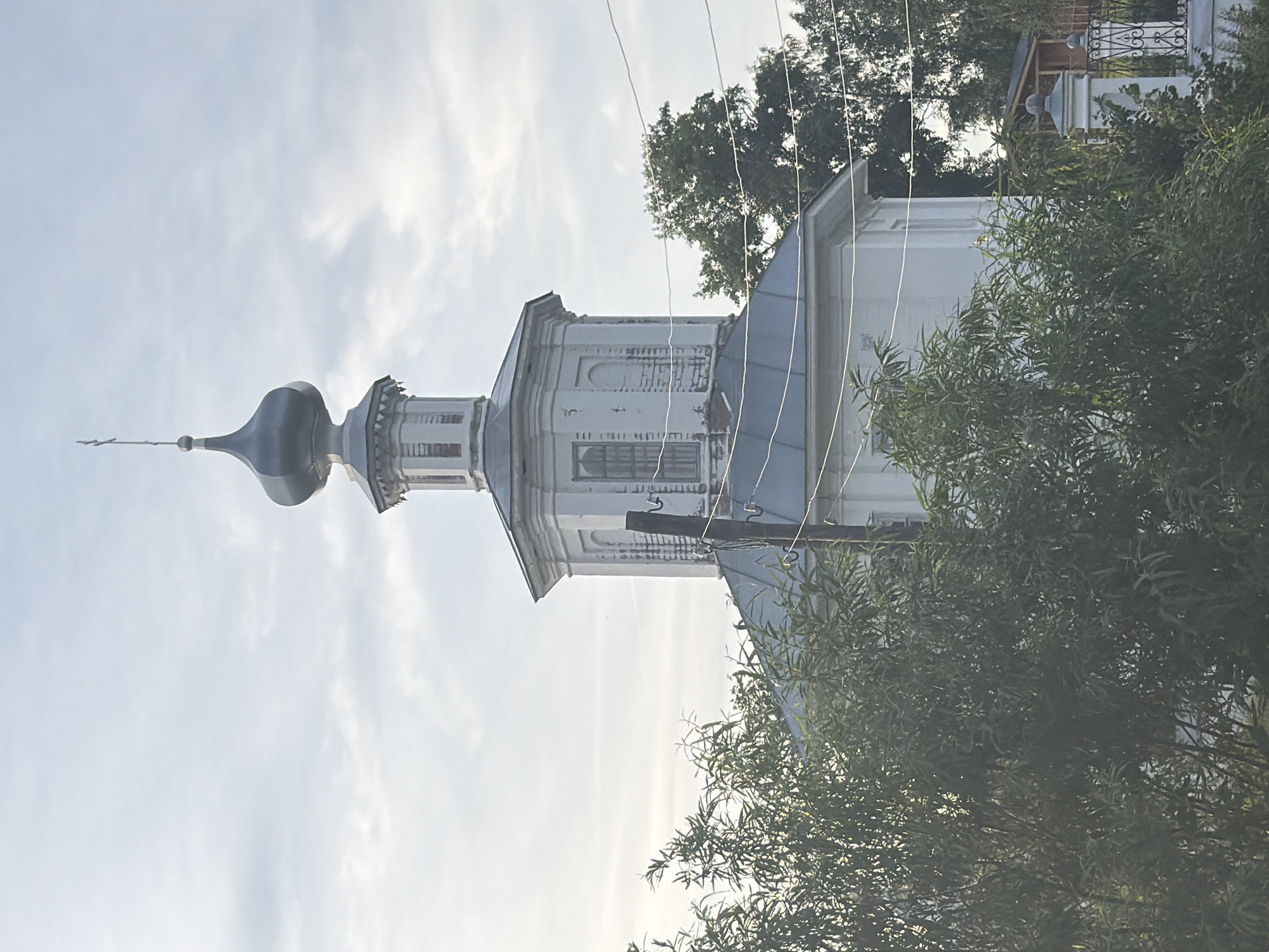 The image shows the bell tower of the Church of the Resurrection in Cherdyn, Russia. The church is white with a gray roof and a dark blue dome topped with a cross. The bell tower is octagonal and has white walls with some visible brickwork. There are windows on each side of the tower. Trees and foliage obscure the lower portion of the church and bell tower. Power lines run horizontally across the image in front of the bell tower. The sky is overcast with some light clouds.