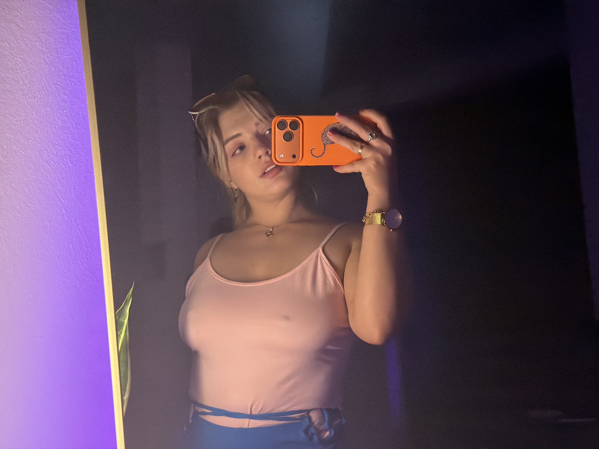 The scene is located in the city of Большая Шильна, Russia. An adult female is captured in a mirror selfie. She has blonde hair pulled back, with sunglasses resting on her head. Her gaze is directed slightly upward and to the right of the camera. She wears a light pink spaghetti-strap tank top, a light-colored necklace with a small pendant, and blue bottoms with visible waist straps. A gold-toned wristwatch with a purple face is on her left wrist, and multiple rings are visible on her right hand. She holds a smartphone encased in a bright orange cover featuring a black triple-camera module and a stylized black leopard print. The indoor environment is dimly lit. A vertical section of a wall or panel illuminated with purple light is visible on the left, separated by a thin vertical yellow strip from a darker background. A single green plant leaf is partially visible behind this yellow strip.