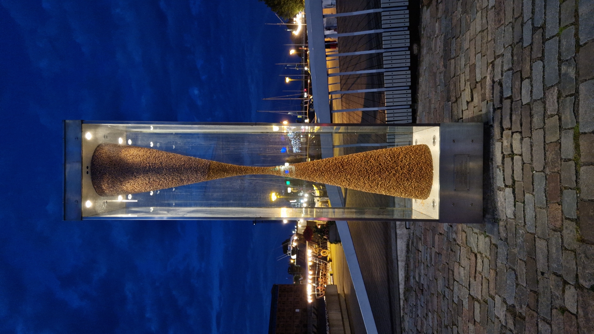 The image depicts an outdoor sculpture, an hourglass filled with what appears to be pebbles or small stones, enclosed within a tall glass and metal casing. The sculpture is illuminated from within by spotlights at the top and a base light. It stands on a paved area constructed of dark, irregularly shaped stones, likely cobblestones, in a city setting. The background shows a waterfront scene at dusk, with a body of water reflecting lights from boats and buildings. Several sailboats are visible docked in the water, along with streetlights and building lights in the distance. A modern-looking railing runs along a wooden deck behind the sculpture. The sky is a deep blue with visible clouds, indicating twilight or early evening. The sculpture is located at the Liepaja Seaside Park, specifically near the former Liepaja Sea Fortress.
