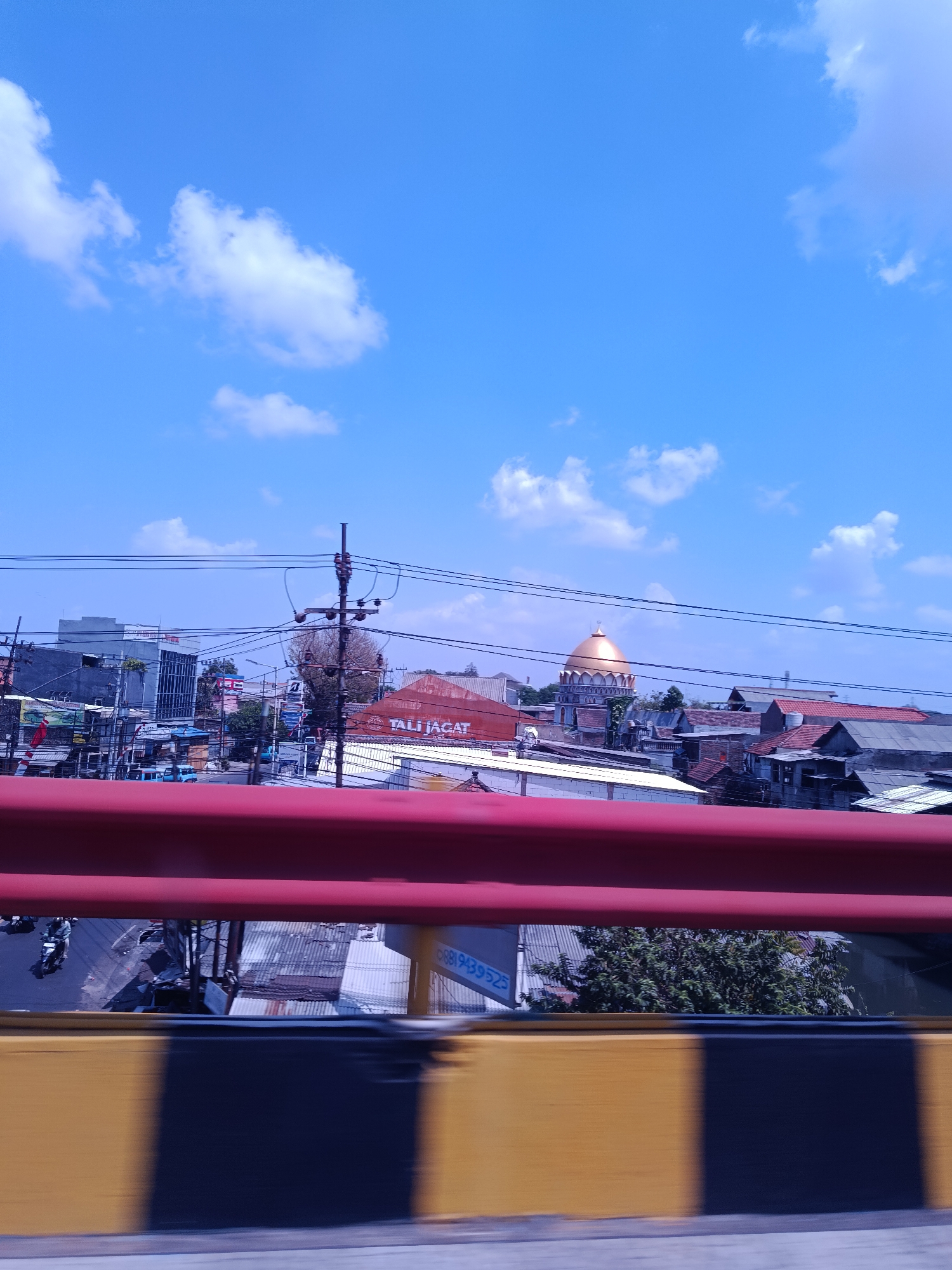The image shows a view of Kedungrejo, Indonesia, captured from a moving vehicle. The foreground is dominated by a red guardrail, with a yellow and black striped barrier below it, indicating the perspective is from an elevated road or bridge. Beyond the barrier, a street scene unfolds under a bright blue sky dotted with white clouds.
Buildings of various sizes and architectural styles line the street, including what appears to be a modern building with blue-tinted windows on the left. Further in the distance, a distinctive golden dome of a building, likely a mosque or a similar religious structure, is visible. Power lines crisscross the frame, typical of urban environments in Indonesia.
On the street level, a few motorcycles are visible, suggesting traffic. Signs, some with text that is difficult to decipher in detail, are present on some of the buildings. The overall impression is of a bustling urban landscape under a clear sky.