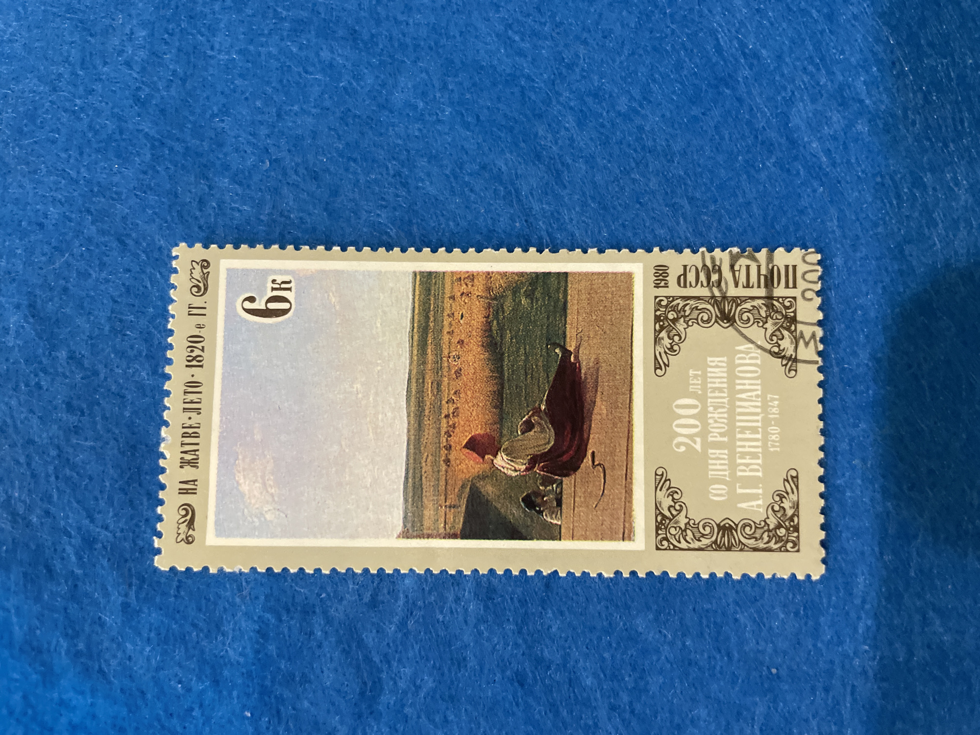 This is a postage stamp featuring a painting of a rural scene. The stamp has a perforated border and is colored in muted tones of brown, yellow, and blue. At the top of the stamp, there is text in Cyrillic, "НА ЖАТВЕ ЛЕТО 1820 г." which translates to "At the Harvest Summer 1820." To the right of this text, a large numeral "6" is present, likely representing the denomination of the stamp.
The central image depicts a woman in a red headscarf and a dark red skirt sitting on a wooden platform, facing away from the viewer. She appears to be resting or watching something. To her left and slightly behind her, another figure, possibly a child, is partially visible. The background shows a golden field of crops, possibly wheat, stretching towards the horizon. A dirt path or road leads into the field, and a few scattered figures can be seen in the distance within the field. The sky above is a light blue with scattered clouds.
Below the painting, more text in Cyrillic is present, indicating "200 лет со дня рождения А.Г. Венецианова 1780-1847." This translates to "200 years since the birth of A.G. Venetsianov 1780-1847," identifying the painting as a tribute to the artist Venetsianov. The year "1980" is also visible, indicating the year the stamp was issued. On the right side of the stamp, vertically oriented text reads "ПОЧТА СССР," meaning "Postage USSR."
The stamp is displayed against a textured blue background, which appears to be fabric, possibly felt or a similar material.
There is no information in the image to suggest it is located in the city of Perm, Russia. The image is a postage stamp and its content is a painting.