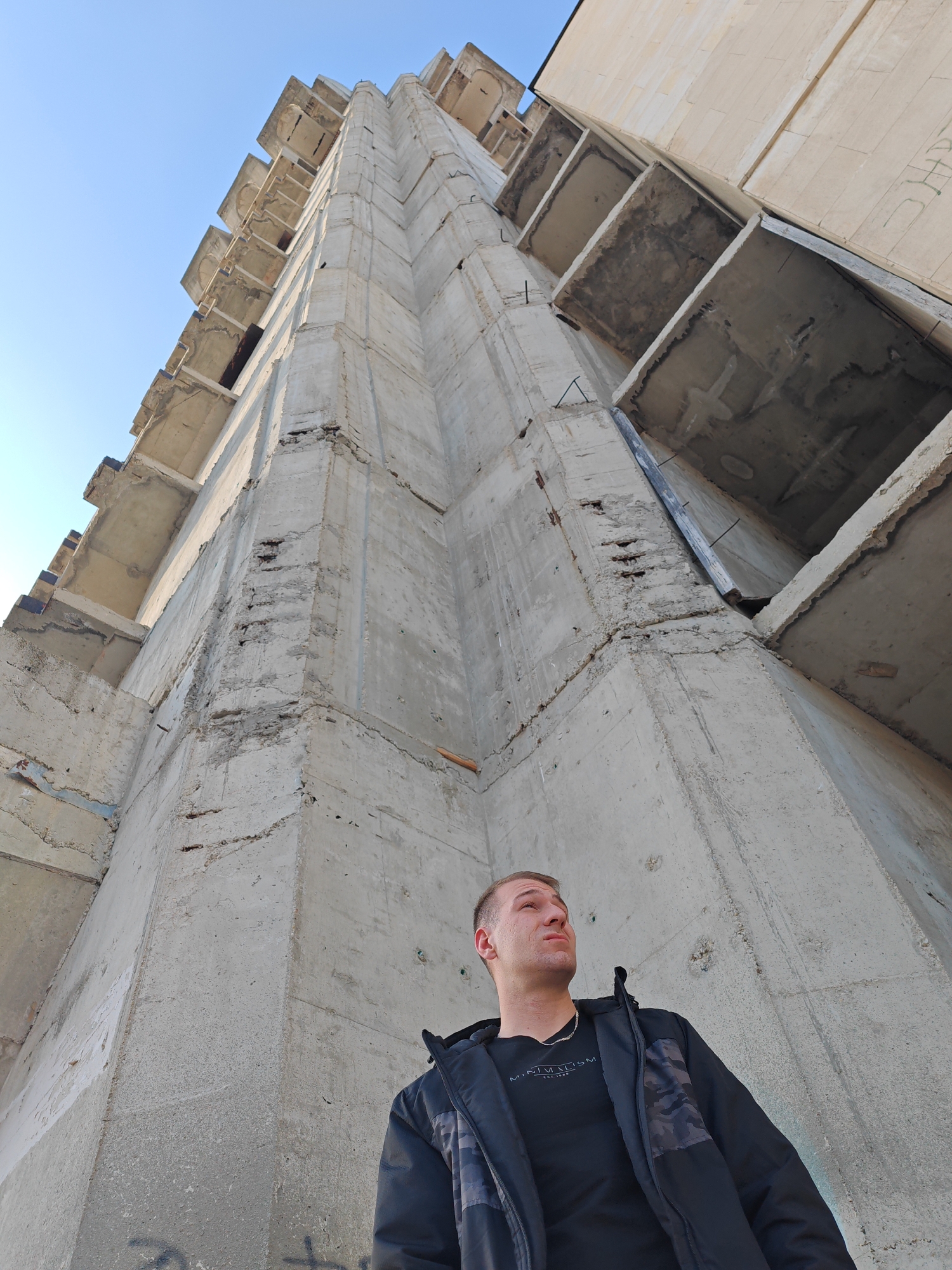 An upward-angled photograph captures a male subject positioned against a towering, unfinished or derelict concrete high-rise building, identified as being in Shumen, Bulgaria. The structure dominates the upper two-thirds of the frame, showcasing raw grey concrete surfaces with visible signs of weathering, erosion, and exposed rebar, indicative of decay or incomplete construction. Numerous open, empty window or balcony apertures are present on multiple stories along the building's facade. The sky in the upper left is clear blue.
The male subject, approximately 20-30 years old, with short light-colored hair, occupies the lower-center of the image. He is wearing a black t-shirt with "MINIMALISM EST. 1999" printed in white on the chest, and an open black zip-up jacket featuring dark grey camouflage panels on the shoulders and upper arms. A silver chain is visible around his neck. His head is tilted slightly upwards, and his gaze is directed towards the upper levels of the concrete building. Faint graffiti is visible on the concrete wall near the bottom left of the frame.