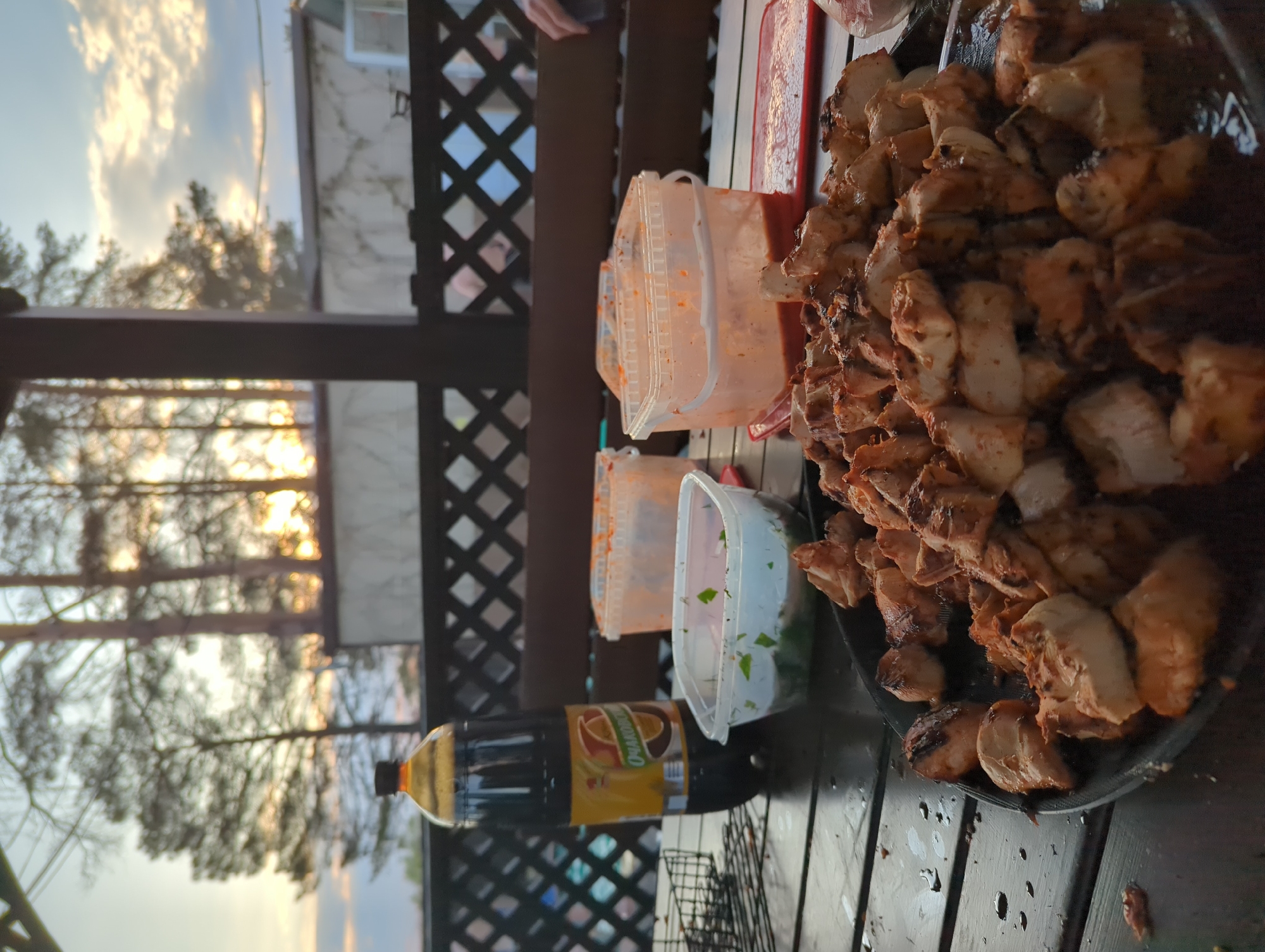 **Scene:**
A horizontal-format photograph taken outdoors, seemingly in the evening, shows a table laden with food and condiments. The scene is situated in the city of Chaikovsky, Russia.
**Objects:**
* A large platter of cubed, cooked meat (likely shashlik) takes prominence on the right side of the table.
* Several clear plastic containers filled with what appears to be sauces and chopped herbs are arranged behind the meat.
* A large bottle of Tarhun-flavored carbonated beverage is positioned near the containers.
* A metal grill, possibly used for cooking the meat, is visible.
**Environment:**
* The table is made of dark-stained wood.
* A lattice-patterned wooden structure forms the backdrop, obscuring the distant view of trees and sky.
* The light from the setting sun peeks through the trees.
**People:**
* Part of a person's arm is visible in the background, suggesting someone is present but not fully in the frame.
* The presence of the food and drinks implies a gathering or meal is taking place.