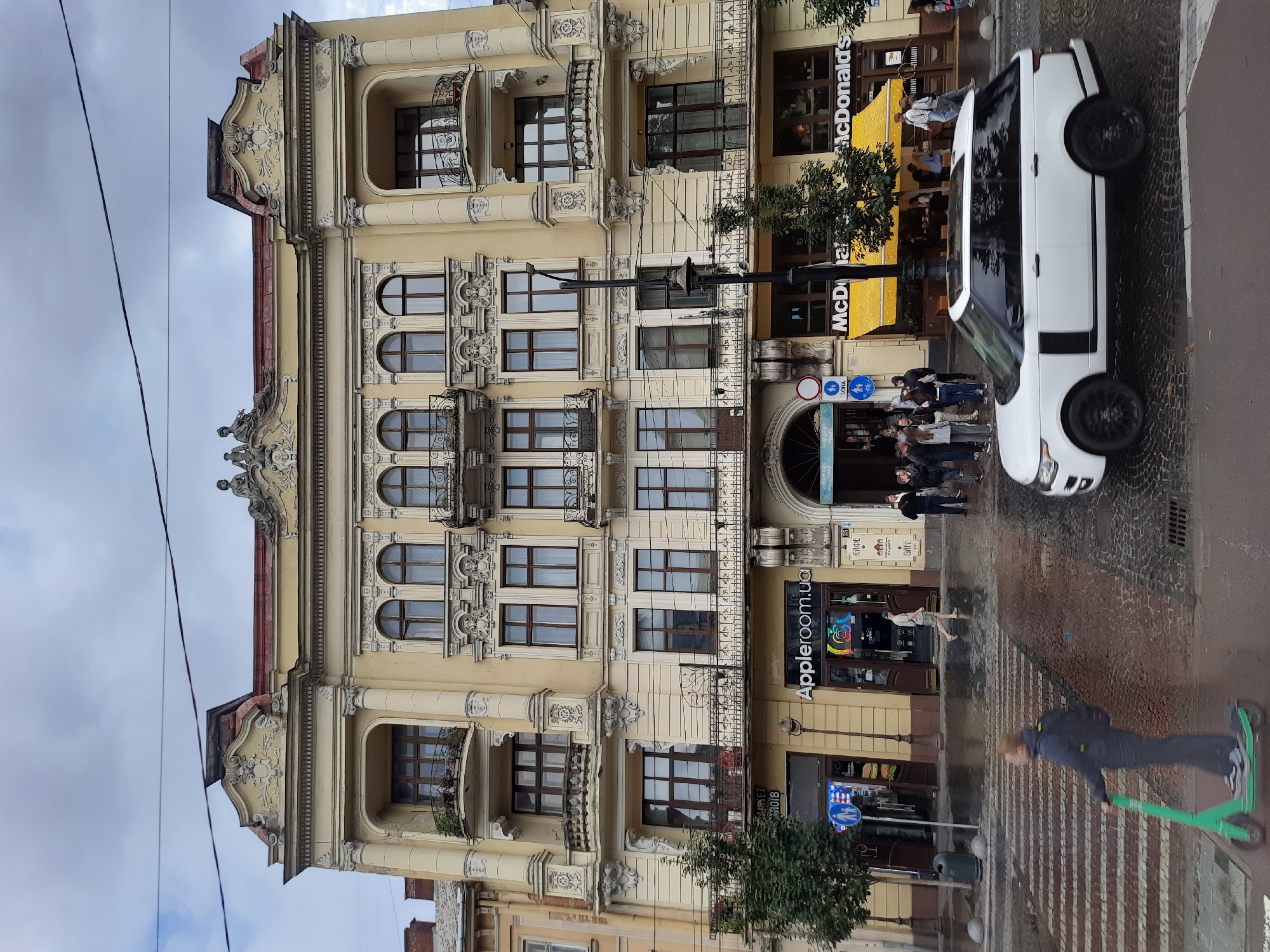 The image shows a street scene in Lviv, Ukraine, on a cloudy day. In the foreground, a person is riding a green electric scooter on a pedestrian crossing. To the right, a white SUV is driving on the cobblestone street. A large, ornate yellow building with several stories stands in the background. The building features decorative architectural elements, balconies with wrought-iron railings, and arched windows. On the ground floor of the building, there are several storefronts, including "Apple room.ua" and "McDonald's," indicated by prominent signage. People are visible walking on the sidewalk and entering the establishments. Street lamps and trees are also present along the street. The overall impression is of a bustling urban environment.