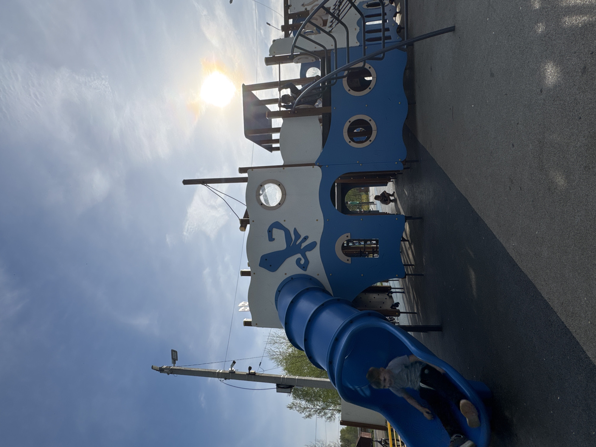 A photograph taken outdoors shows a playground structure, resembling a ship. The structure is primarily blue and white, with a blue tube slide extending from one end. A child is sliding down the tube slide. The playground is located in Shevlyakovo, Russia. The sky is blue with the sun shining brightly, causing a lens flare effect in the image. A utility pole with electrical wiring is visible to the left of the slide. The playground surface is paved. There are other indistinct figures visible on the structure.