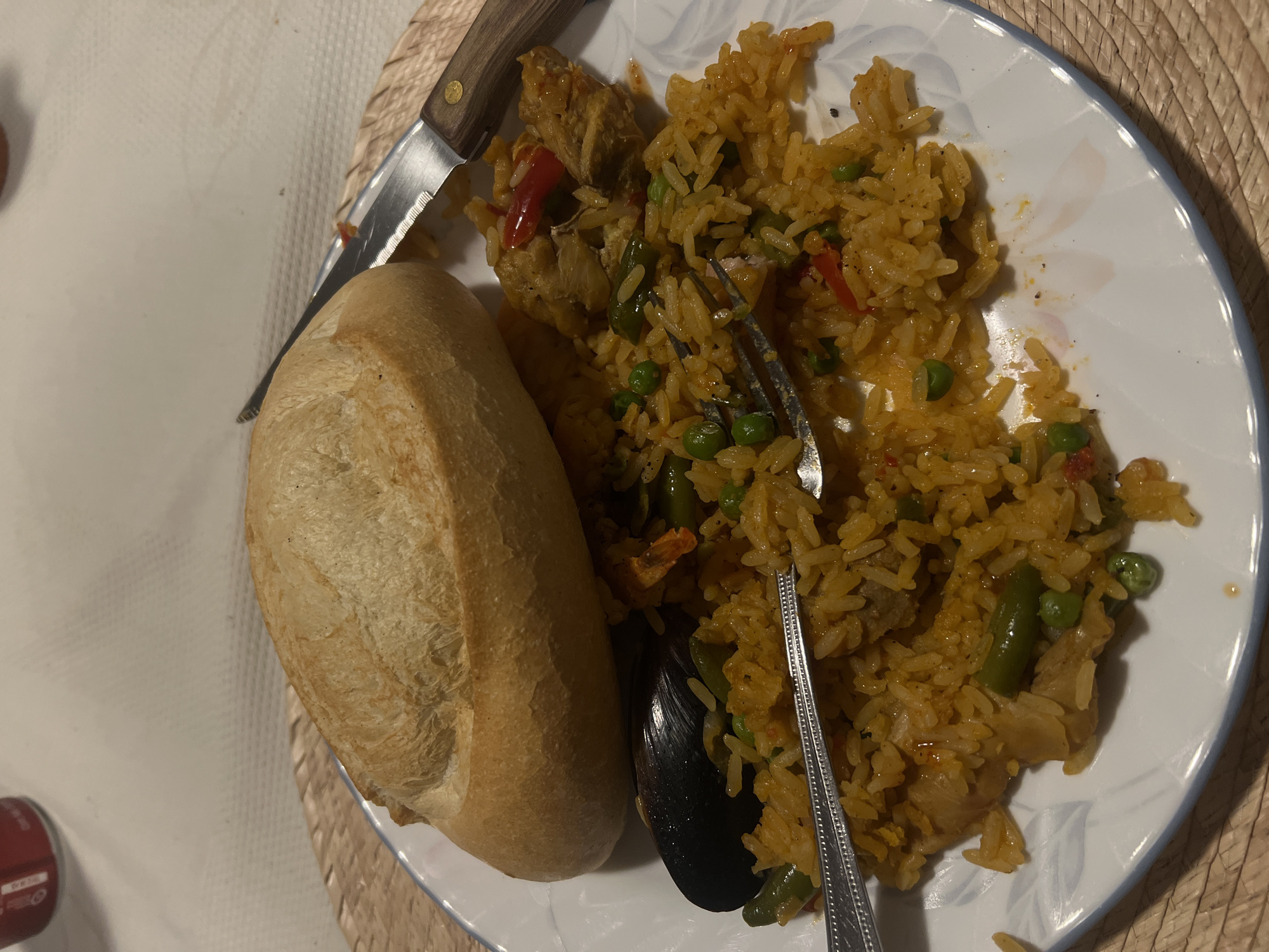 On a white plate with a blue rim, sits a serving of paella. The paella includes rice, peas, green beans, mussels, red bell peppers, and what appears to be chicken. A fork is partially buried in the rice. A bread roll is next to the paella, and a knife rests on top of the bread. The plate sits on a woven placemat on a white tablecloth. A small red can is visible in the lower left corner. There are no visible clues indicating the location is in Mexico City.