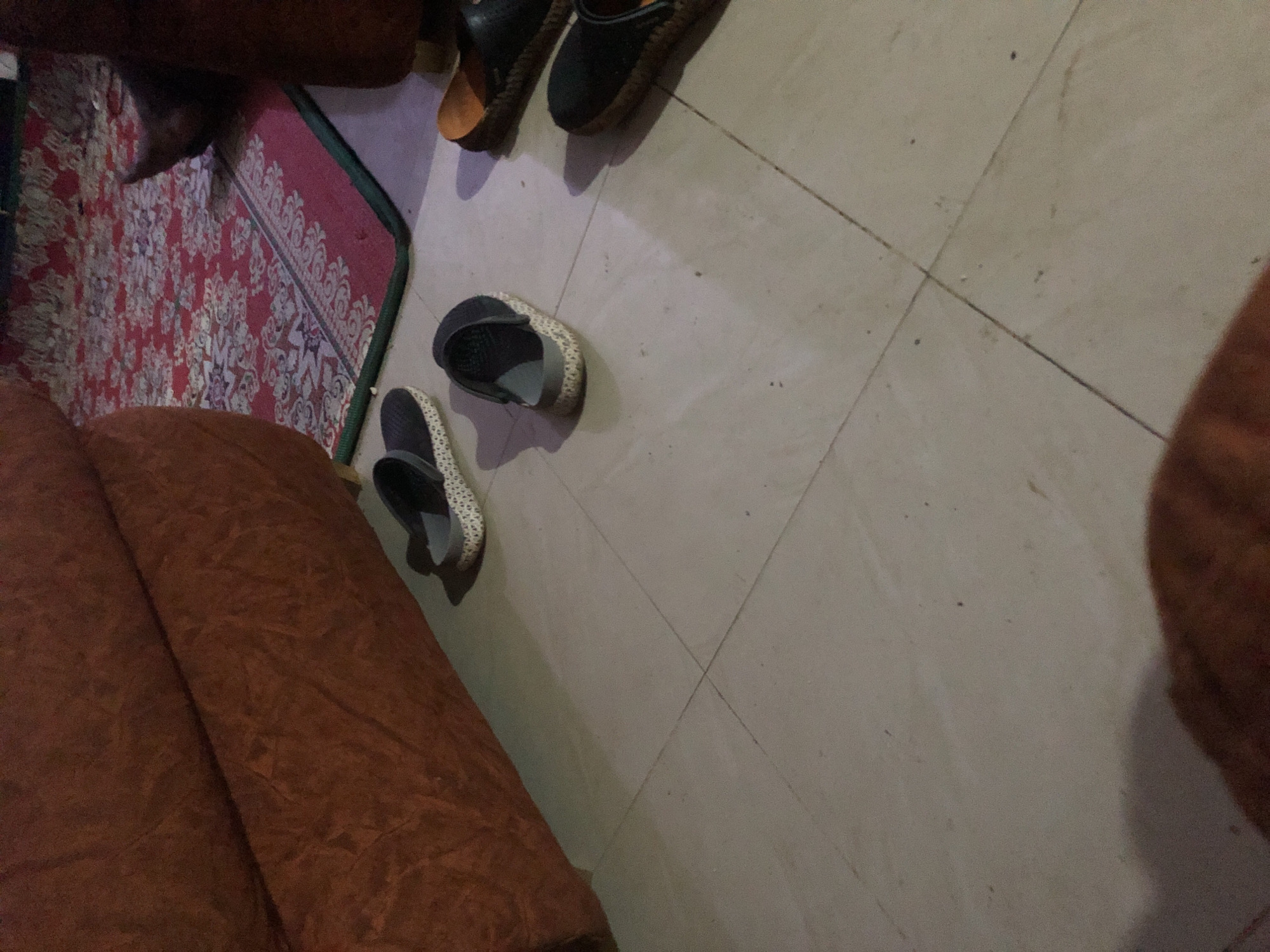 The image depicts a floor scene in what appears to be a residential setting in Kabong, Nigeria. The floor is tiled with light-colored square tiles, with visible grout lines. Several pairs of footwear are scattered on the floor. To the left, a portion of a reddish-brown sofa is visible. In the background, a red rug with a patterned design is laid out on the floor, partially covered by a dark object, possibly another piece of furniture. The lighting suggests an indoor environment, possibly with artificial light.
Specifically, there are at least four pairs of shoes visible:
- One pair of dark gray clogs with a textured white sole is on the left side of the image, near the sofa.
- Another pair of similar dark gray clogs, with the back strap down, is positioned slightly to the right and center.
- Further to the right, there are two pairs of black sandals or flip-flops with brown soles. One is slightly more visible, showing a brown sole and black straps. The other is partially obscured.