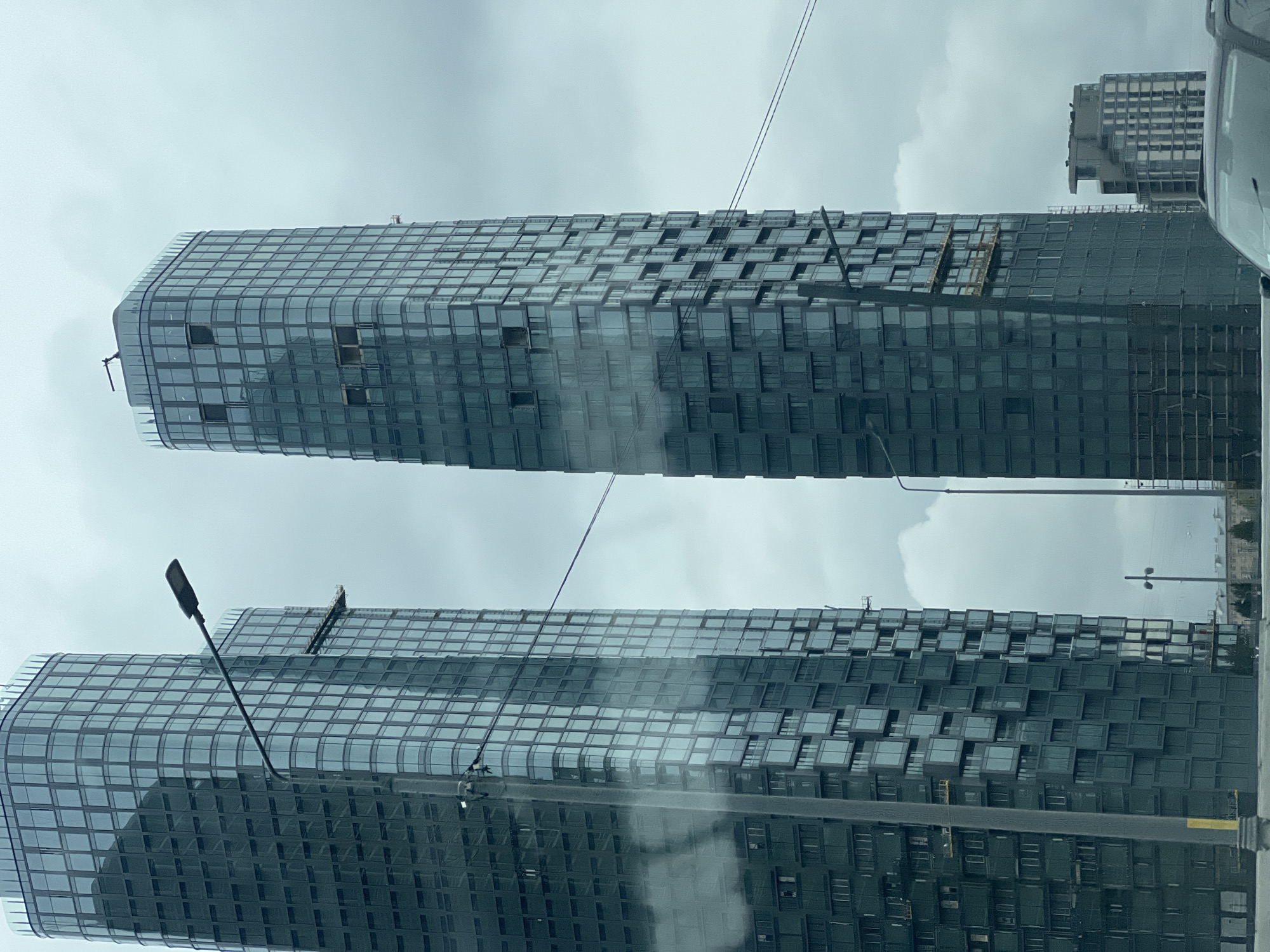 The image shows two tall buildings in Moscow, Russia, viewed from a lower vantage point. The buildings are modern, high-rise structures with glass facades arranged in a grid-like pattern, with protruding window/balcony elements. Some windows on the buildings are missing or open. There is visible construction or maintenance work occurring on the buildings, with scaffolding present. A third building is visible in the background to the upper right. An overcast sky is visible. Light posts are in the foreground, further obscuring the view of the buildings. The image appears to have been taken from inside a vehicle.