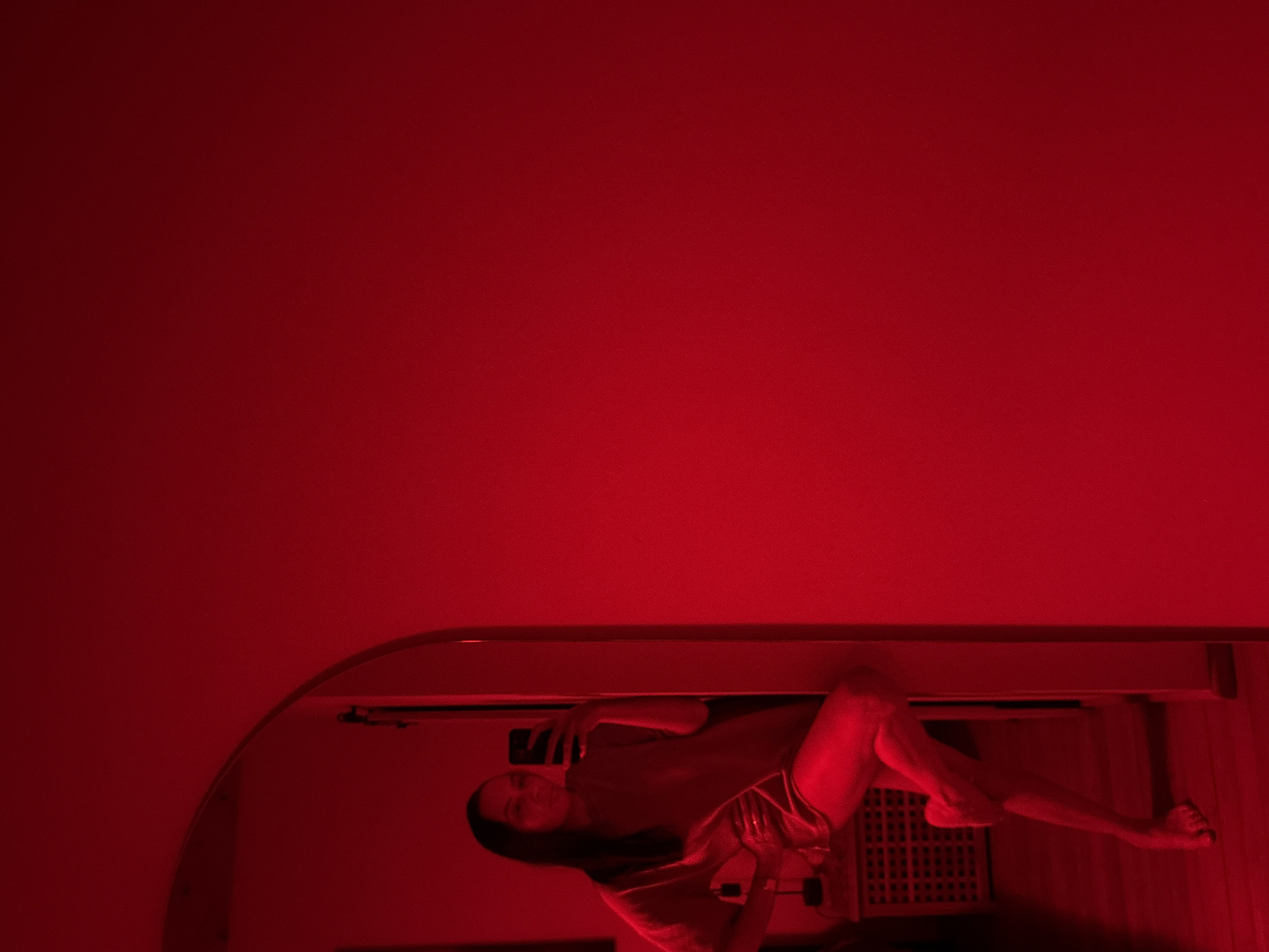 The image displays an indoor scene illuminated by intense red lighting. A female individual is captured in a mirror's reflection, positioned on the left side of the frame. She has dark hair and is wearing a loose-fitting, possibly oversized, t-shirt or dress, with her legs exposed. Her left hand rests on her hip, while her right hand holds a dark-colored smartphone, indicating a selfie-capture interaction with the mirror. She is barefoot, with her left leg bent and crossed over her right leg, leaning against a doorframe or wall. Visible in the reflected background are a low, slatted piece of furniture (possibly a cabinet or radiator cover) and a lamp. The flooring appears to be wood. The right two-thirds of the image consists of a plain wall, uniformly bathed in red light. The specific location within the city of Chaikovsky, Russia, is not ascertainable from the visual evidence.