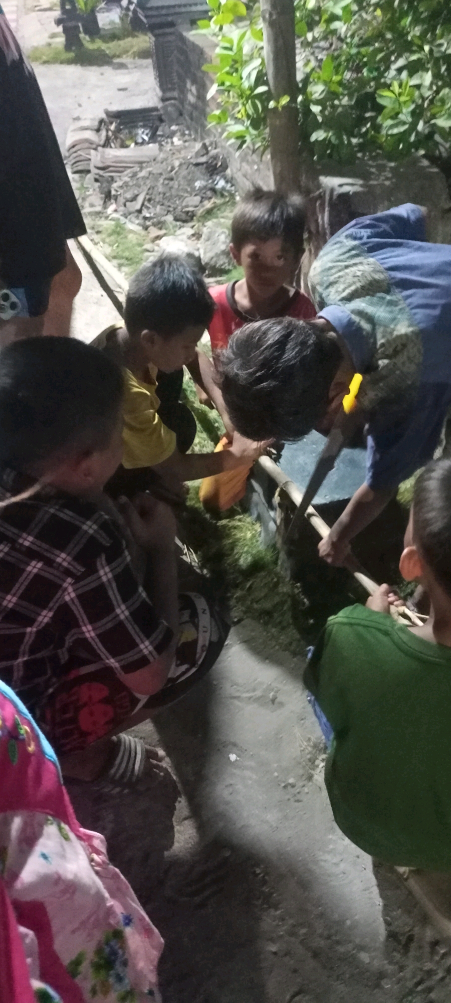 In Gempolklutuk, Indonesia, a group of children are gathered around a hole in the ground, appearing to be digging or removing something from it. The ground is a dirt and concrete mix, with some scattered debris and green foliage visible. The children are dressed in casual clothing, including a plaid shirt, a yellow t-shirt, and a red t-shirt. One child is wearing a blue shirt and appears to be assisting with the digging using a shovel. Another child, wearing a green shirt, is also positioned near the hole, potentially observing or participating. The scene is dimly lit, suggesting it may be dusk or night. A black backpack with white lettering is visible on the ground near one of the children.