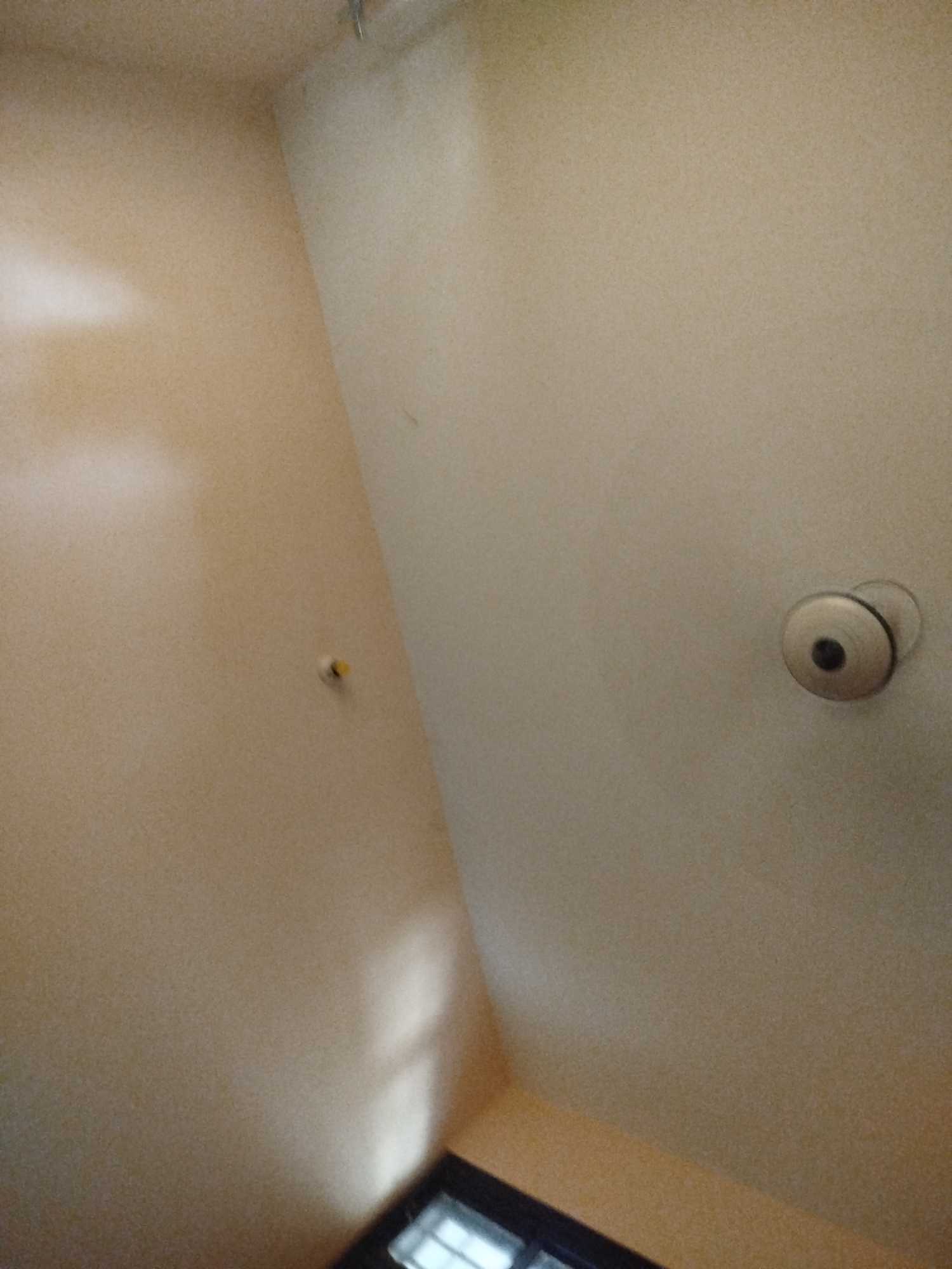 A security camera is mounted on the wall in the corner of a room. The camera is pointing at a wall with a yellow circular object. A window is visible in the lower right corner of the image.