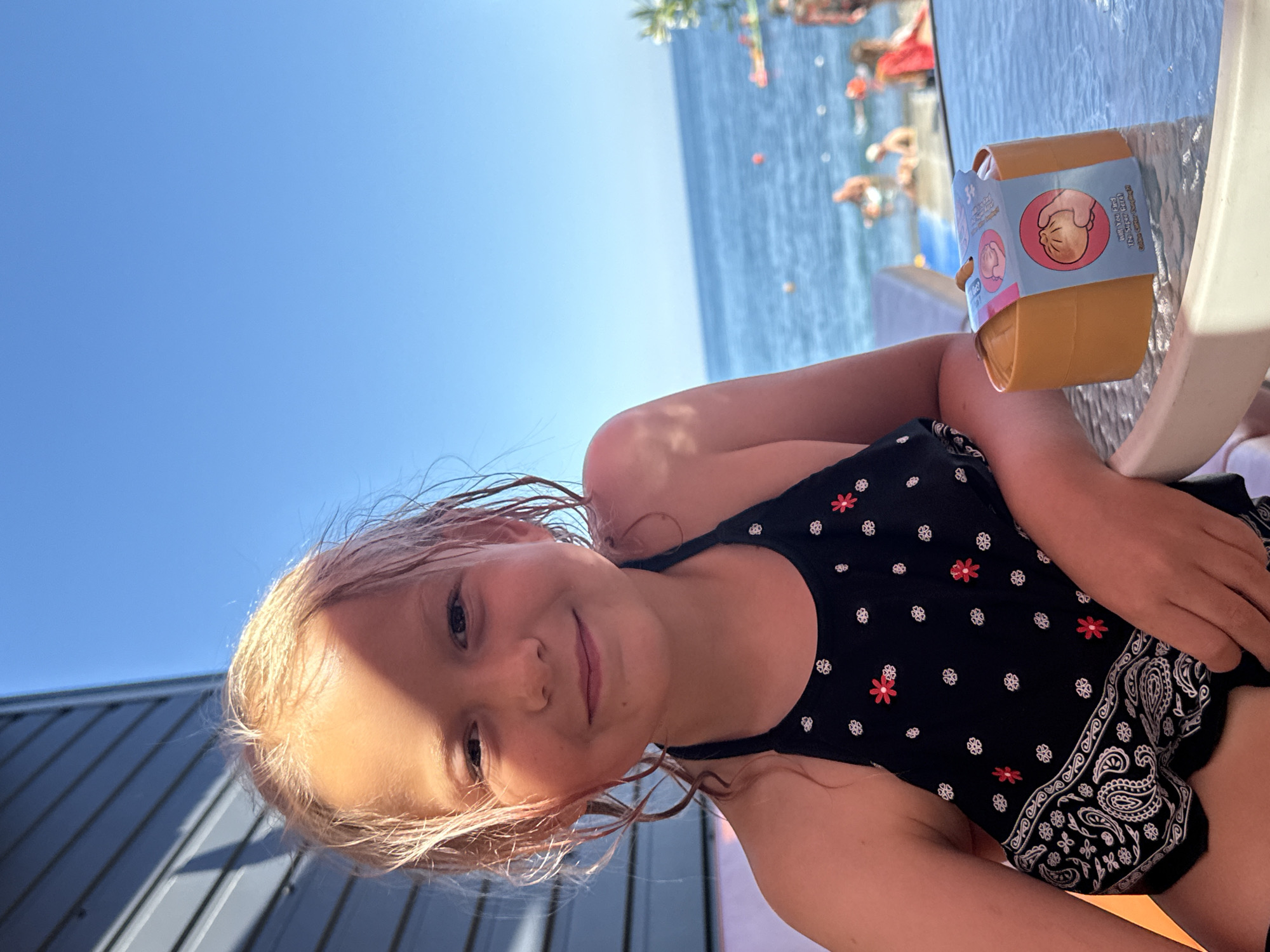 A young girl with blonde, wet hair smiles at the camera, her face illuminated by the sun. She is wearing a black swimsuit with a white floral pattern and a paisley trim. The image is taken outdoors on a sunny day, with a bright blue sky visible in the background. Behind the girl, a body of water, likely the sea, stretches out to the horizon. In the background, there are blurry figures of people enjoying themselves near the water, suggesting a beach or poolside setting. On a table beside the girl is a small, brown toy container with a blue label. The label features an illustration of a hand holding a round object and some text, including the number "3+" and what appears to be Ukrainian or Russian script. The overall mood of the image is cheerful and relaxed, indicative of a holiday or a day spent enjoying the outdoors. The corrugated siding of a building is visible on the left side of the frame.