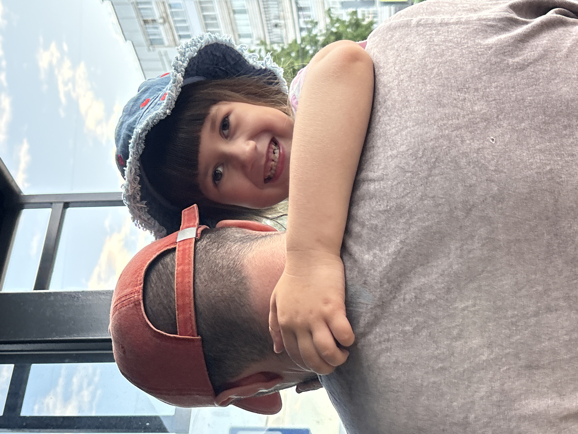 The image, captured in the city of Chaikovsky, Russia, presents a close-up, over-the-shoulder view of an adult from behind, with a young child interacting with the adult and facing the camera. The adult, appearing male, wears a faded orange baseball cap reversed, featuring an adjustable strap with a silver-toned buckle. His garment is a light gray t-shirt, exhibiting a textured fabric and a small imperfection on the upper back. The child, approximately 4-7 years old, has dark hair with bangs and wears a blue denim-style bucket hat adorned with red polka dots and a distinctly frayed brim. Her expression is a joyful, wide smile, revealing a gap from a missing upper front tooth. The child's right arm is wrapped around the adult's neck and rests on his shoulder, indicating an affectionate embrace. The background, viewed through large, dark-framed windows, shows a bright blue sky with scattered white clouds, alongside distant multi-story urban buildings and green tree foliage, typical of an outdoor city environment under natural daylight.