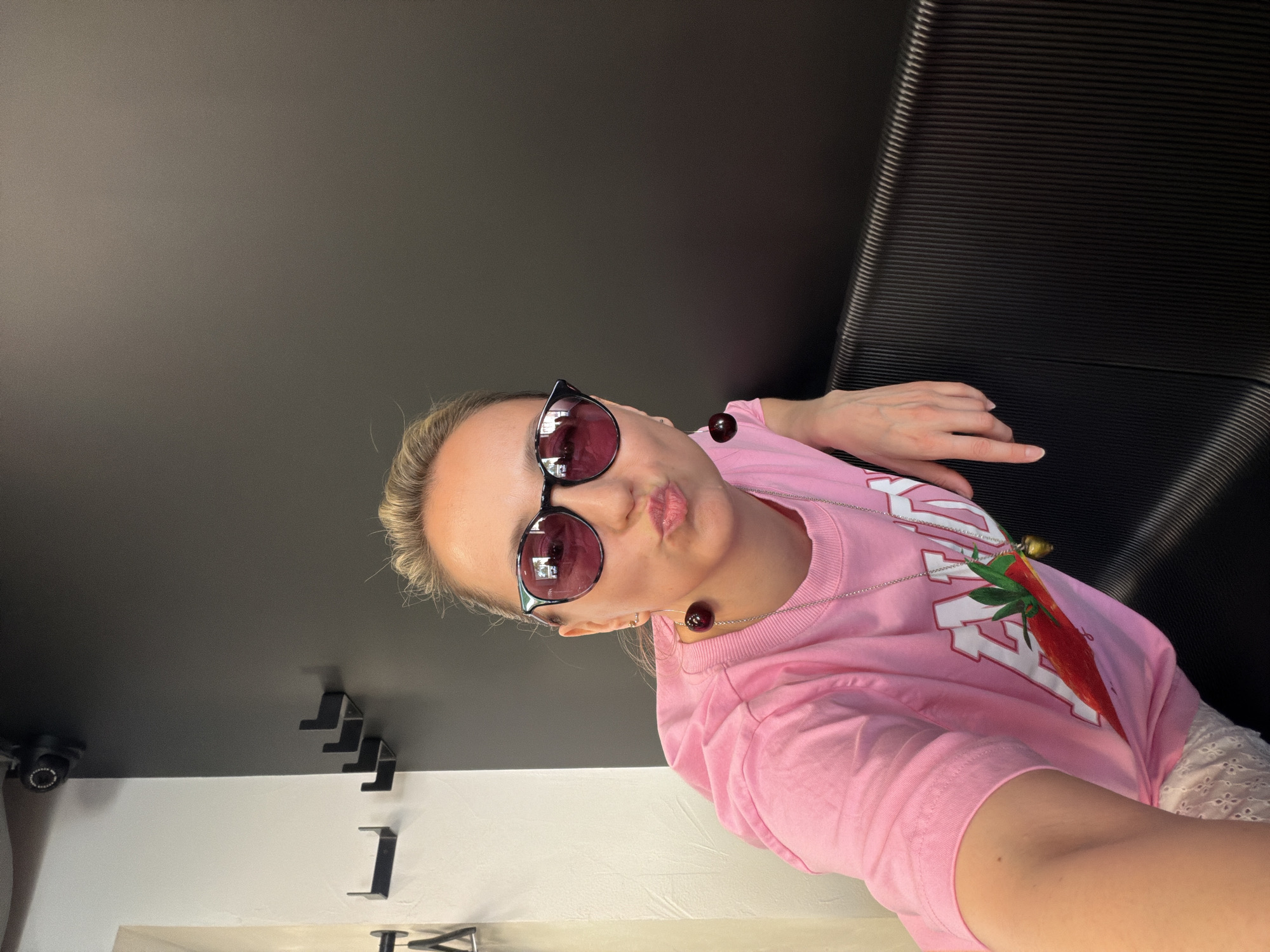 A young woman with sunglasses and a pink strawberry-themed shirt is in Izhevsk, Russia. She is in a room with a dark ceiling and white walls. A security camera is mounted on the ceiling. A black bench is behind her. She is looking at the camera with pursed lips. She has two cherries, each attached to a cord, around her neck.