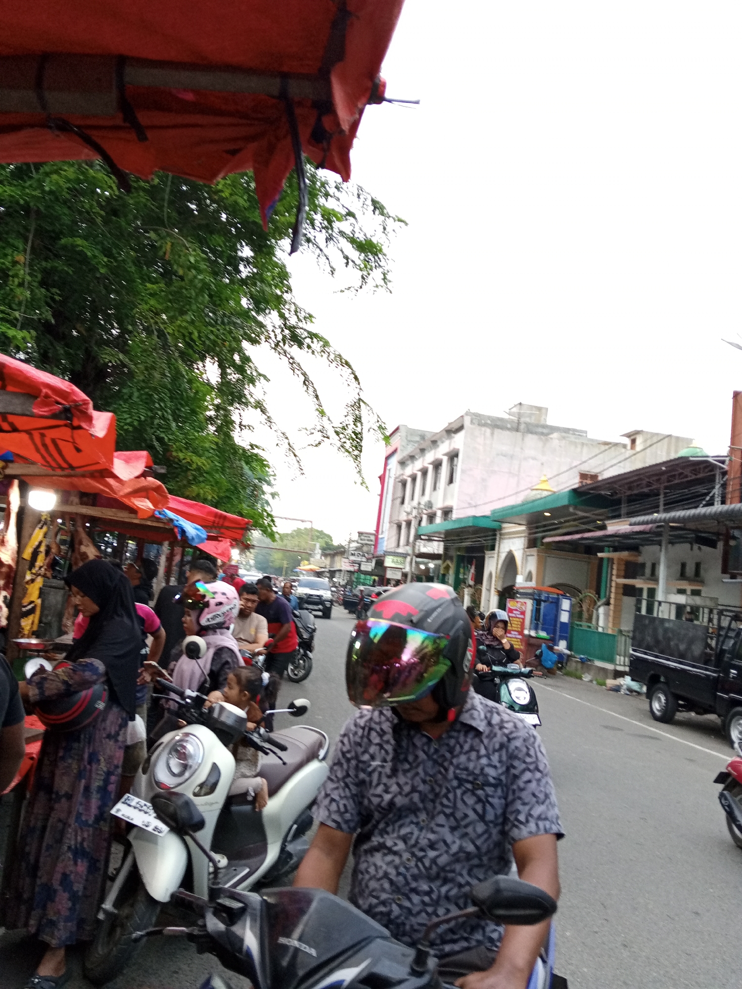 The image captures a busy street scene in Puloara, Indonesia. Several people are on motorcycles, some wearing helmets. One motorcycle has a small child seated on the front. A group of people is gathered around a vendor stall, recognizable by a red awning, that potentially sells meat. Several cars and trucks are on the road, and there are buildings along the street. The sky is overcast.