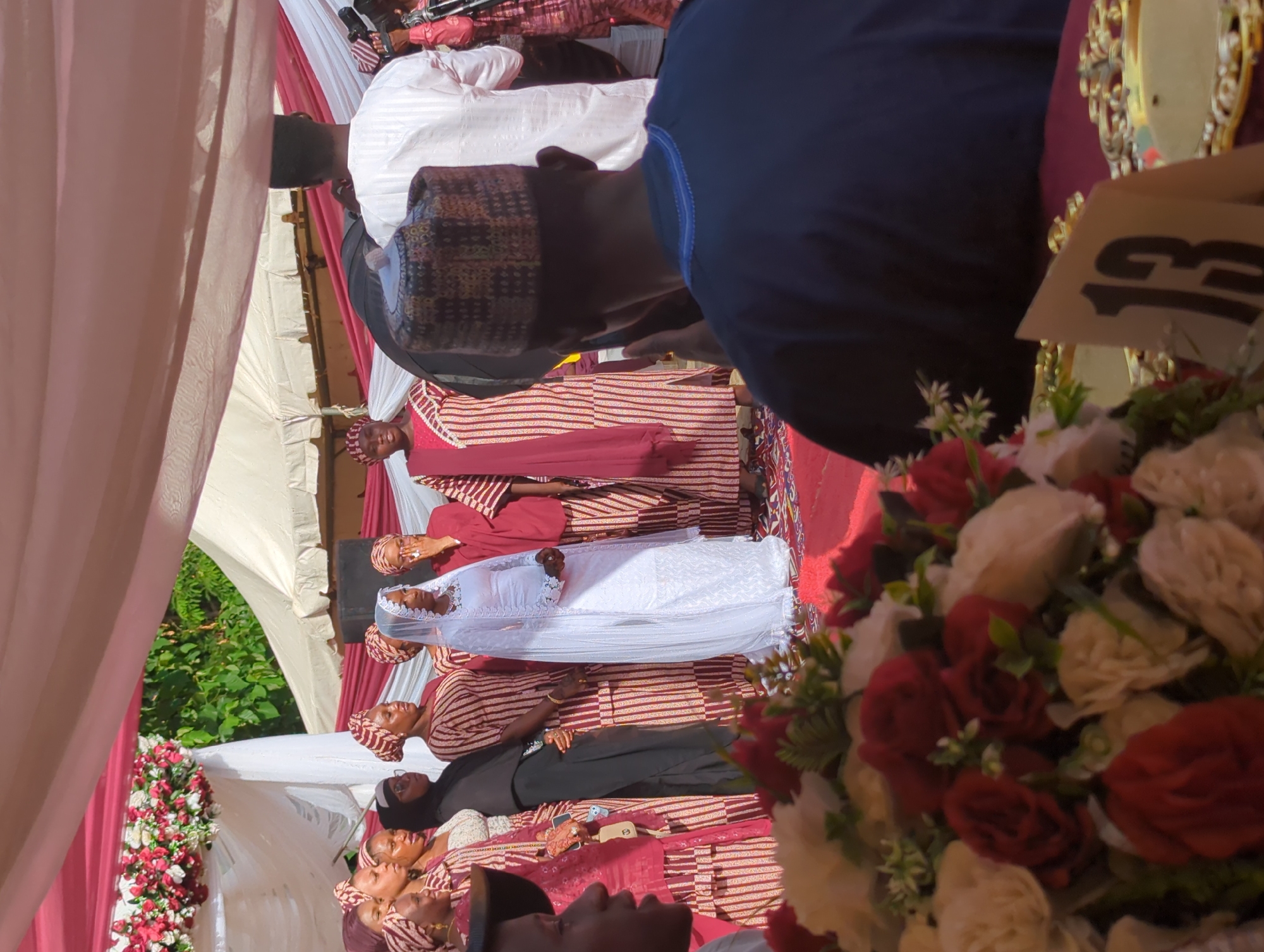 The image captures an outdoor daytime celebration in Kabala, Nigeria, under a large fabric canopy. The scene is observed from a slightly elevated position behind a table. In the immediate foreground, the top of a table covered with a deep red/maroon fabric is visible. A blurred artificial floral arrangement, primarily composed of red and cream-colored roses and other flowers with green foliage, occupies the bottom left. To the right of this arrangement, a white tent card with the number "13" printed in dark numerals is placed. Further to the right, a gold-colored, ornate decorative object is partially visible on the table. On the far right foreground, the back of an individual is visible, wearing a dark blue garment with a light blue embroidered collar trim and a patterned cap in shades of brown, black, and tan. This individual is facing away from the viewer, looking towards the central activity. A partial profile of another person, dark-skinned and wearing a dark cap or hat, is visible on the far left foreground, also looking towards the center. The midground features a group of approximately twelve individuals standing on a raised platform or stage, covered with a red carpet or fabric. The central figure, positioned slightly to the right of the frame's vertical center, is a woman dressed in a floor-length white gown with intricate lace detailing and a sheer white veil cascading down her back. She stands with her hands lightly clasped in front of her. Surrounding her are several women wearing matching traditional attire consisting of vertically striped gowns in maroon/red and cream/white, complemented by matching headwraps. One woman immediately to the bride's right has her right hand raised to her mouth. Another woman, further to the left within the group, wears a dark head covering and a dark, possibly black, abaya, and spectacles. To the far right of the group, the back of a man in a white striped garment is visible, with dark hair, seemingly observing or documenting the event; camera equipment is partially visible to his right. Above the central figures, a large canopy of draped white and reddish-pink fabric stretches across the frame, providing shade. An elaborate floral arrangement, featuring red and white flowers with green foliage, is attached to the canopy structure on the far left. The background reveals natural green foliage, indicative of trees or bushes, and multiple light-colored, pointed tents, suggesting an outdoor event