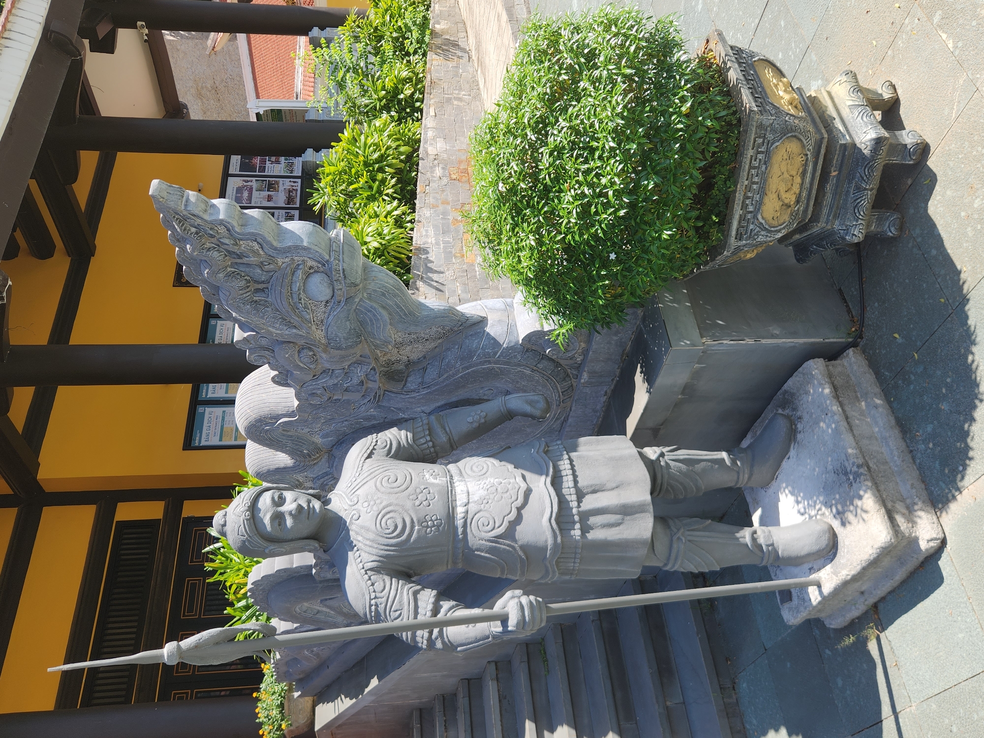 The image captures a stone statue of a warrior holding a spear, standing to the left of a large, intricately carved stone dragon head. The statue is positioned in front of a building with a bright yellow facade and dark brown pillars. Stairs are visible to the left of the warrior statue. To the right of the dragon head, a lush green bush is planted in an ornate, dark gray and gold pot. The scene is outdoors, under a bright sky, with shadows indicating sunlight. The location is identified as the entrance of the Phap Lam Pagoda in Đà Nẵng, Vietnam.