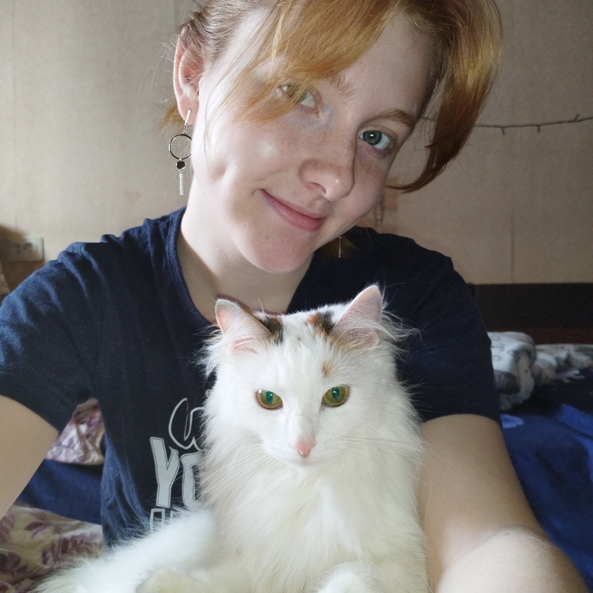 A young woman with reddish-blonde hair and freckles smiles gently at the camera while holding a long-haired white cat. Her light-colored eyes and dimples are visible as she gazes slightly off-camera. She is wearing a dark blue t-shirt with partial white lettering, including "Y" and "O", visible on the front. She has a distinctive dangle earring in her left ear, featuring a hoop, a dark circular element, and a rectangular bar adorned with small embellishments, along with other ear piercings.
The white cat, with patches of brown and orange fur on its head and ears, has striking green-gold eyes and a calm, focused expression. It is positioned in front of the woman, being held close.
The scene is indoors, likely a casual home environment, possibly a bedroom. The background features a plain light brown or beige wall. Hints of bedding or blankets in blue and patterned fabrics are visible behind and to the sides of the subjects. A string of small lights is faintly visible running horizontally across the upper right portion of the background, and an electrical outlet can be seen on the left. The overall impression is one of warmth and affection between the woman and her feline companion.