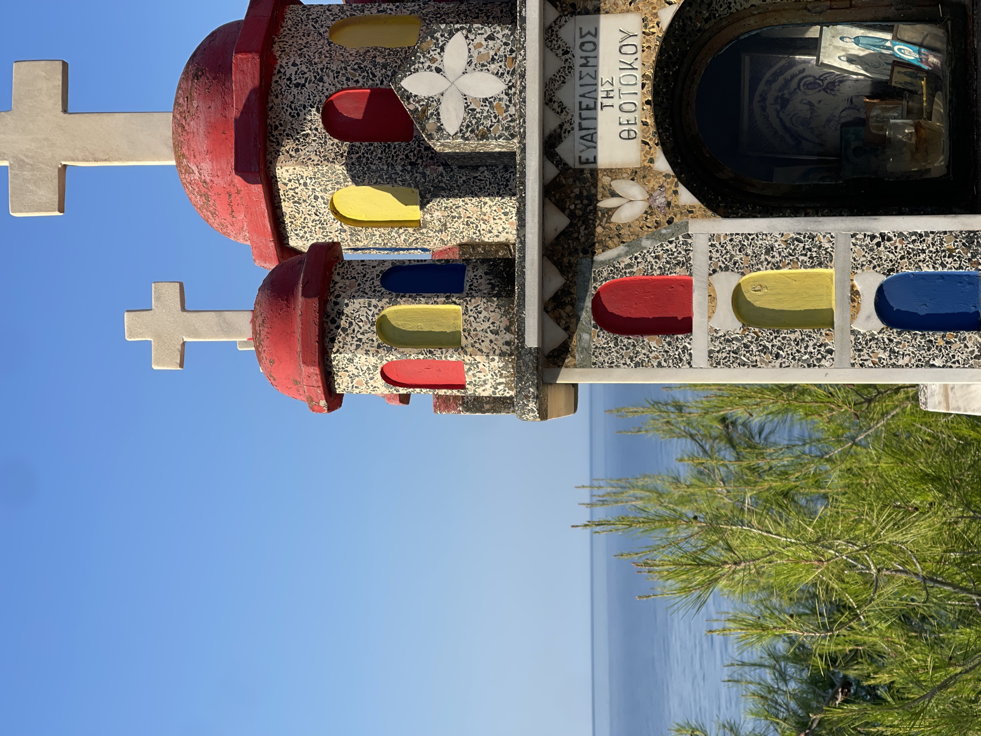 A small, colorful, Byzantine-style church is situated on a cliff overlooking the Aegean Sea in Sithonia Municipal Unit, Greece. The church is made of speckled concrete and adorned with red, yellow, and blue arched windows. Two white crosses top its red domes. Inscriptions in Greek are visible on the church's facade. Pine trees with needle-like leaves frame the foreground. The sea stretches to the horizon under a clear blue sky. The church appears to be a votive offering or a memorial chapel.